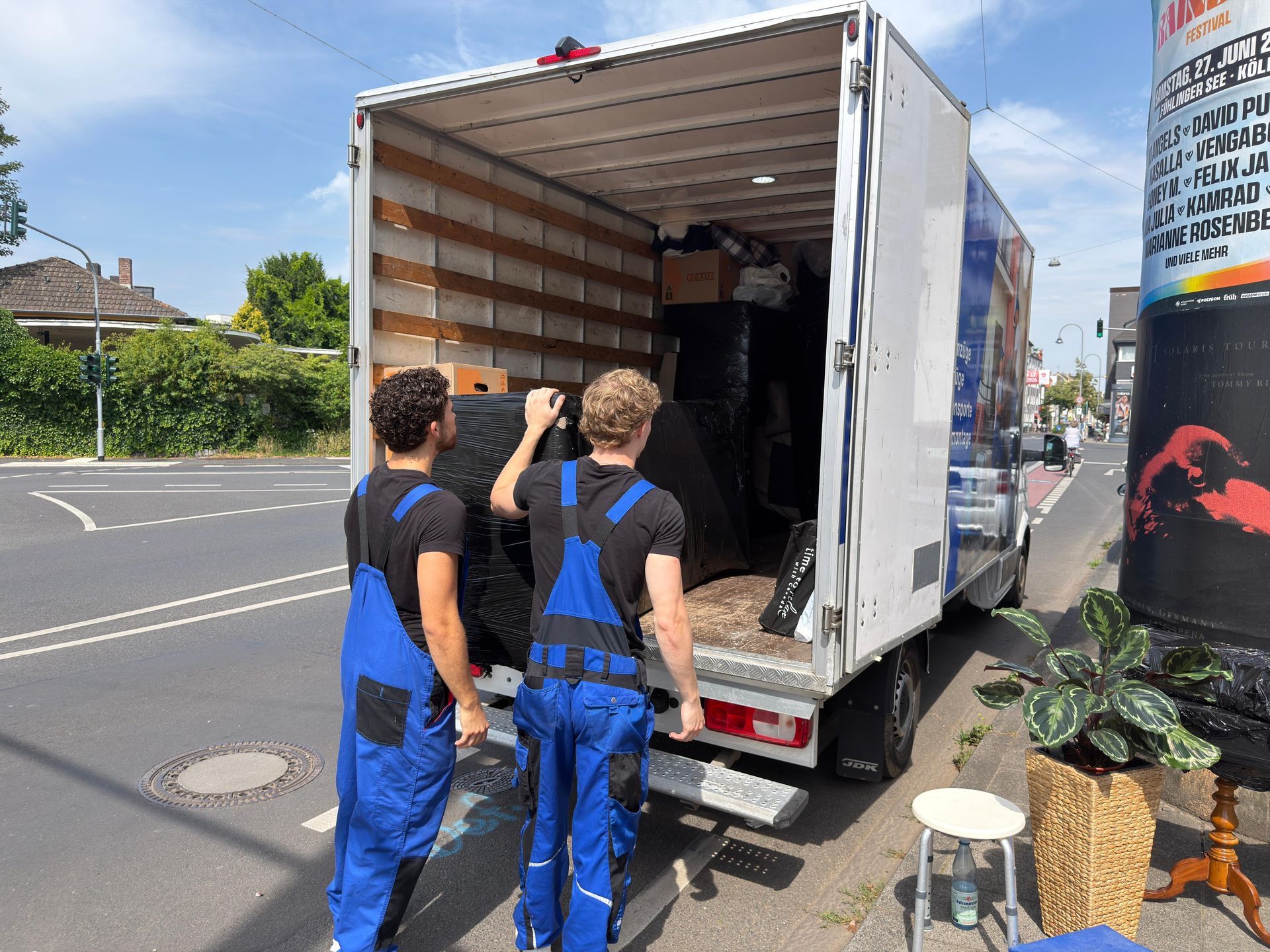 Kisten und Pflanzen in einem Raum, im Hintergrund bewegen sich Umzugshelfer.