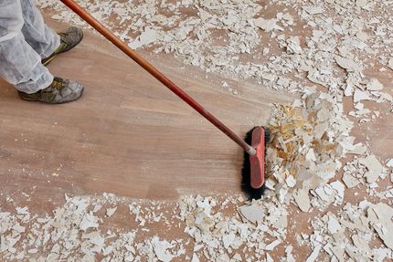 Lars Kaufhold belädt einen Eimer mit Reinigungsmitteln aus einem Lieferwagen. Außenaufnahme am Tag. Mitarbeiter in Schutzanzug fegt Bauschutt von einem Holzboden – Baureinigung in Kerpen
