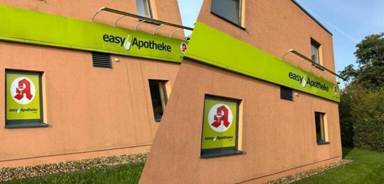 Die Fassade der„Easy Apotheke“. Das Gebäude ist orange mit einem grünen Schild und einem Schaufenster mit Apothekenauslage.