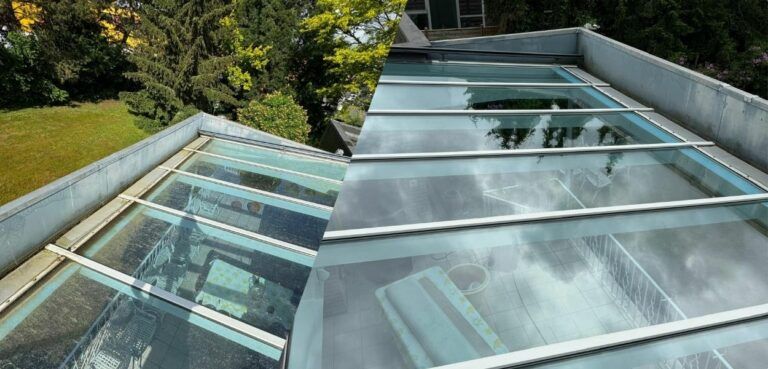 Vorher-nachher-Bild: Glasdach mit Metallrahmen. Im Hintergrund grüne Bäume und Rasen.