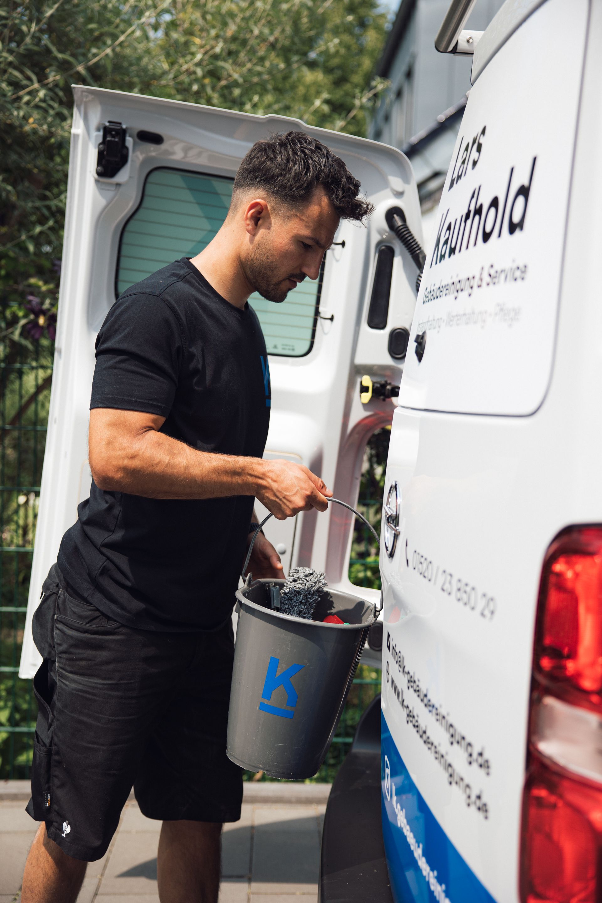 Lars Kaufhold belädt einen Eimer mit Reinigungsmitteln aus einem Lieferwagen. Außenaufnahme am Tag.