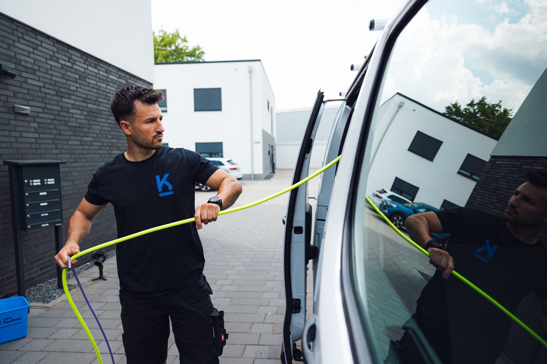 Mann reinigt im Freien, er zieht einen gelben Wasserschlauch aus dem Firmenwagen.