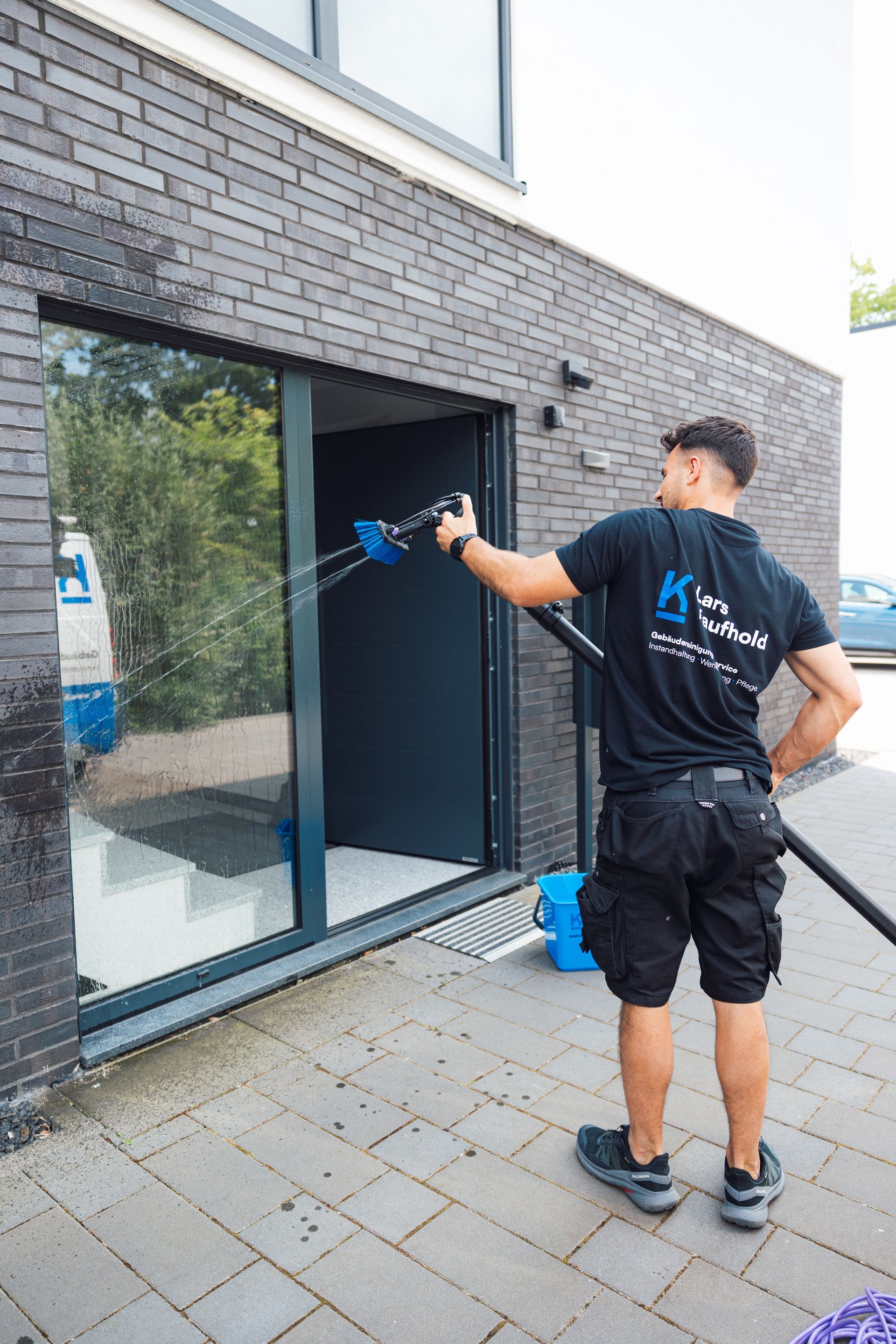 Fensterputzer Lars Kaufhold sprüht eine Glastür ab. Er steht vor einem Gebäude, neben ihm steht ein kleiner blauer Eimer.