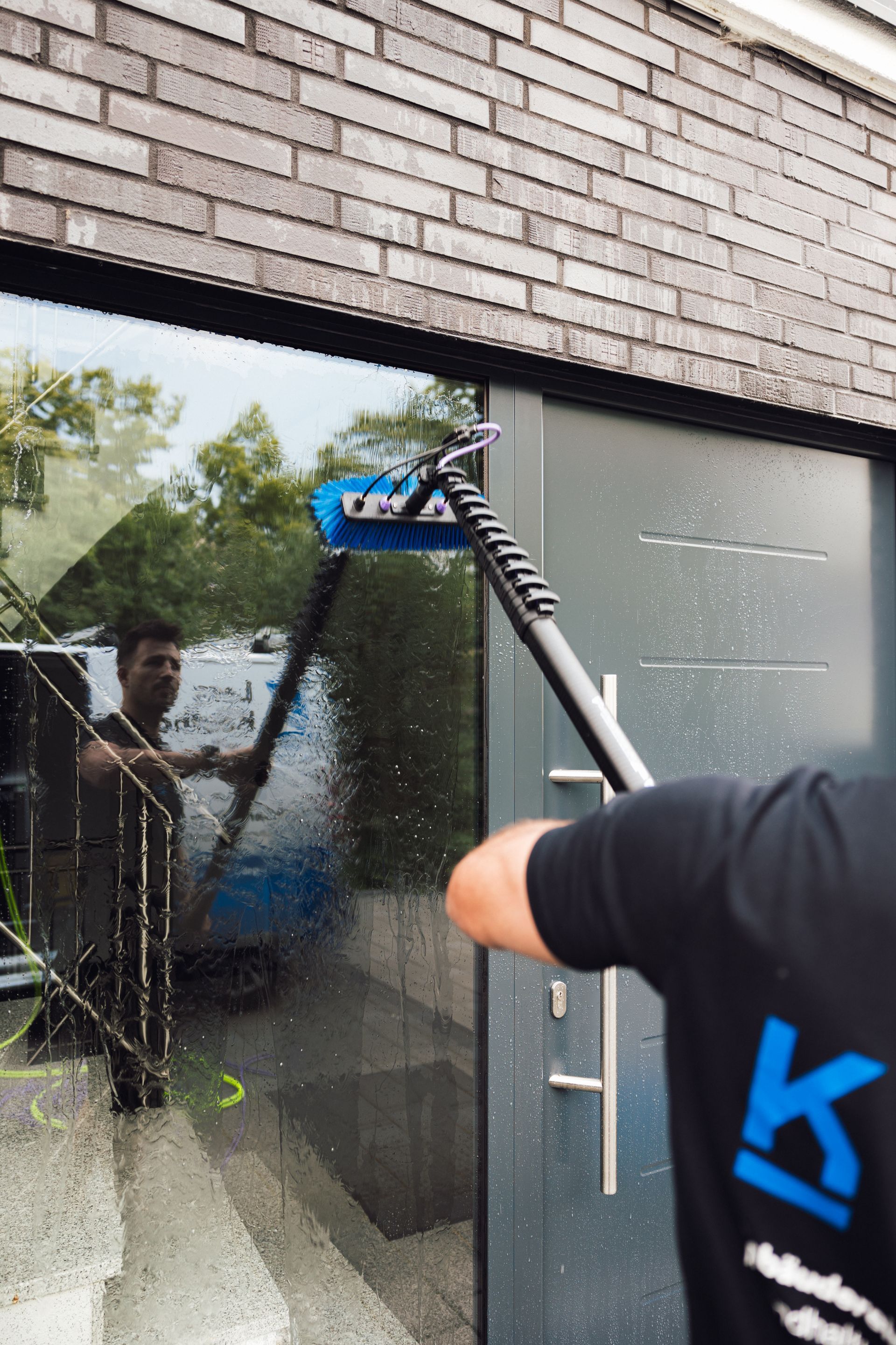 Eine Person putzt mit einem langstieligen Werkzeug ein Fenster. Gebäudeaußenansicht, sonniger Tag.