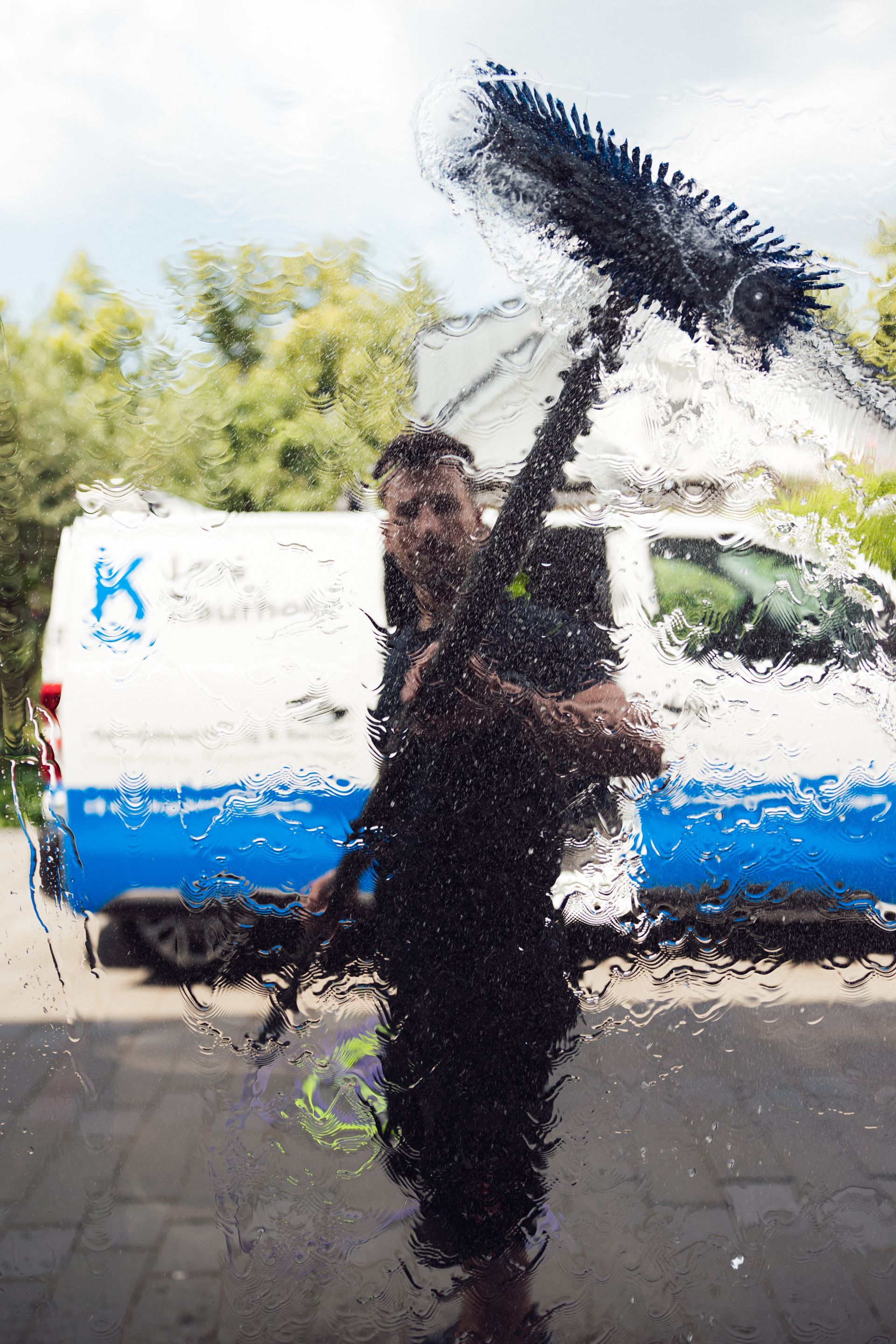 Ein Fensterputzer mit einem langen Reinigungsbesen spiegelt sich im Glas. Im Hintergrund ein weiß-blauer Firmenwagen.