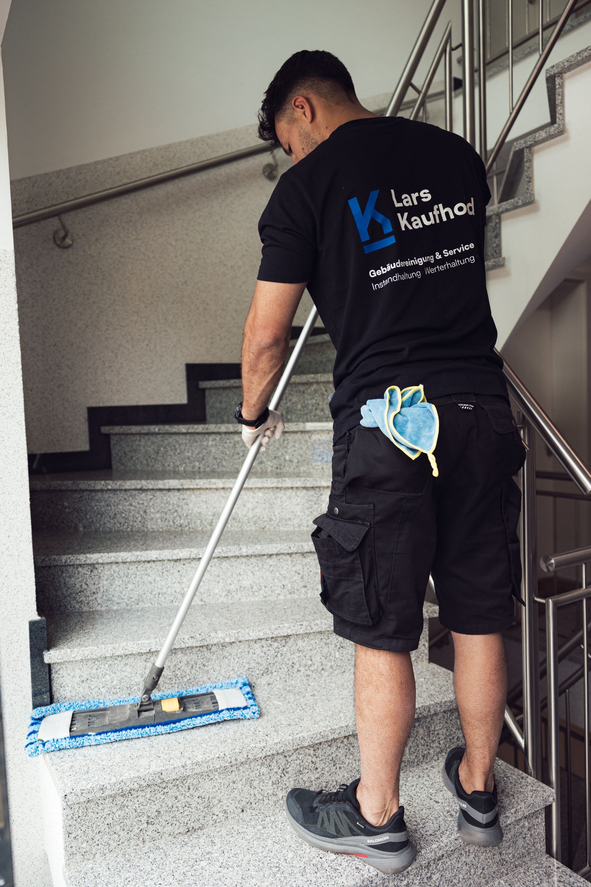 Eine Person wischt die Treppe in einem Gebäude. Er trägt ein schwarzes T-Shirt, Shorts und Turnschuhe.