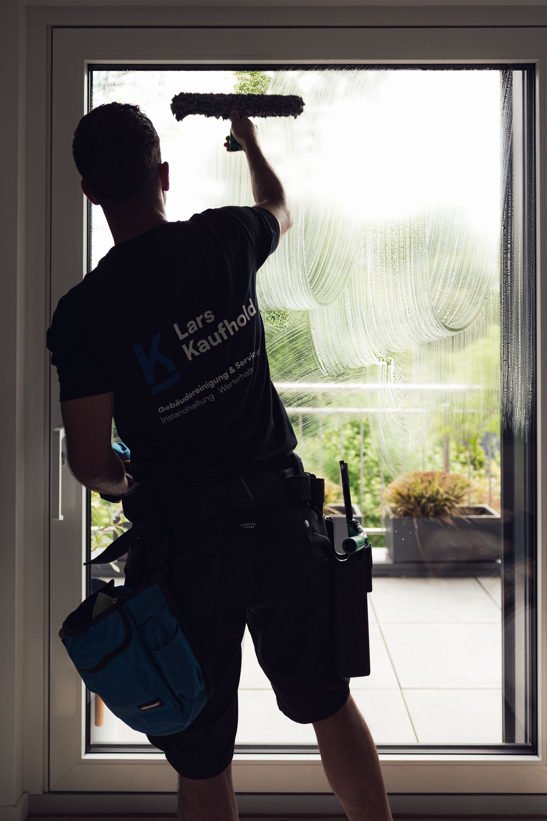 Fensterputzer Lars Kaufhold sprüht eine Glastür ab. Er steht vor einem Gebäude, neben ihm steht ein kleiner blauer Eimer.
