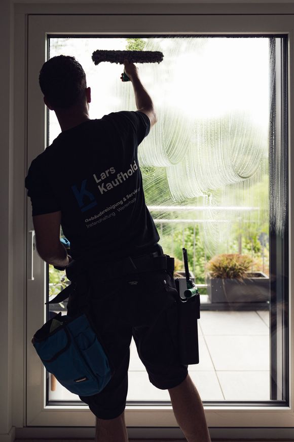 Fensterputzer Lars Kaufhold sprüht eine Glastür ab. Er steht vor einem Gebäude aus grauen Ziegelsteinen, neben ihm steht ein kleiner blauer Eimer. Fensterputzer Lars Kaufhold sprüht eine Glastür ab. Er steht vor einem Gebäude, neben ihm steht ein kleiner blauer Eimer.