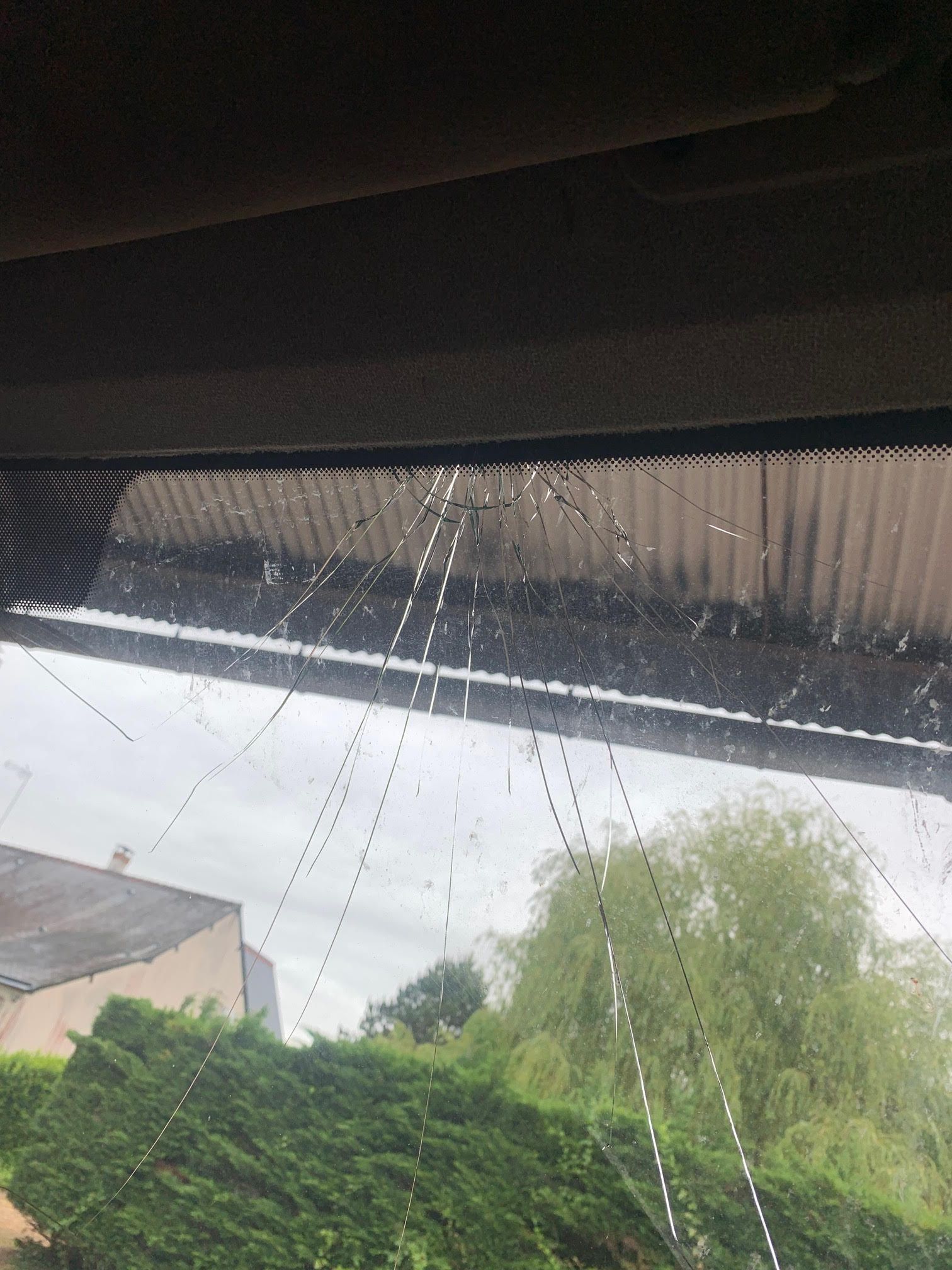 Des traînées de pluie sur la vitre d'une voiture, avec des arbres et une maison à l'extérieur par temps couvert.