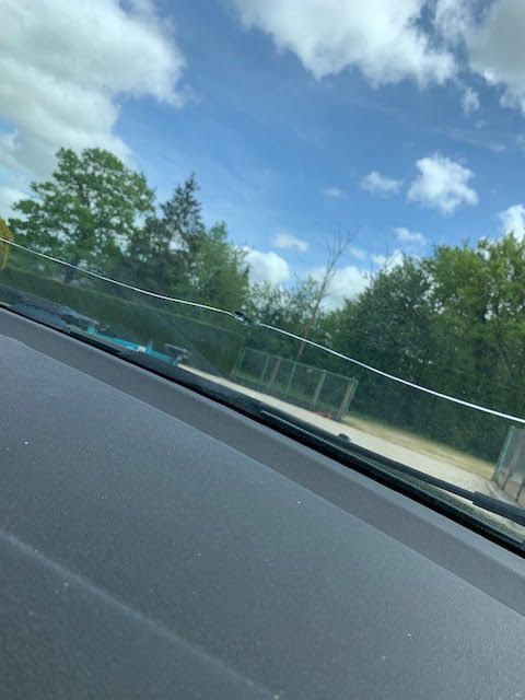 Vue depuis le tableau de bord d'une voiture : une clôture en bord de route, des arbres et un ciel bleu nuageux.