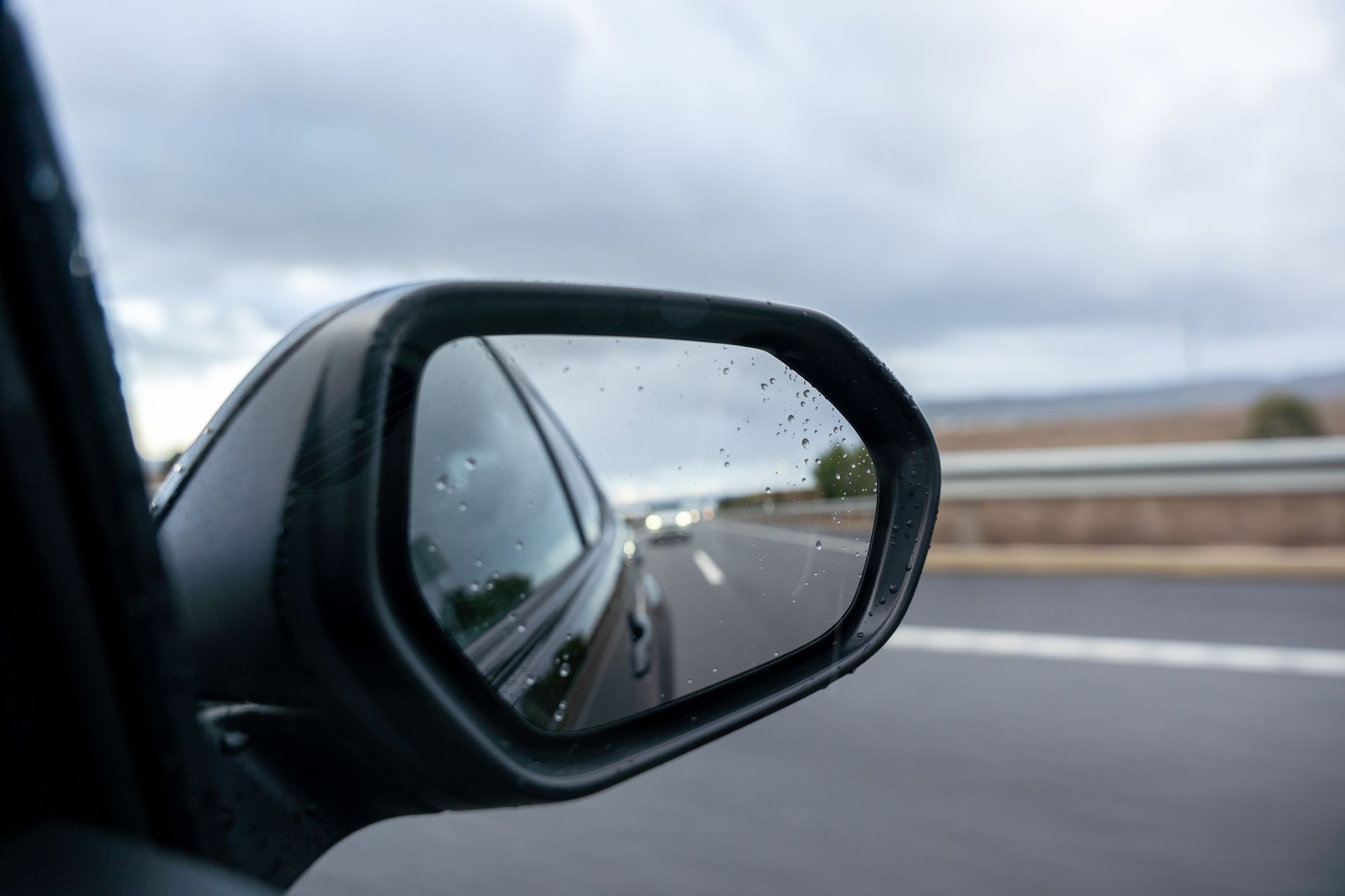 Le rétroviseur latéral d'une voiture reflète une autoroute mouillée sous un ciel nuageux, avec la route et le garde-fou flous sur le côté.