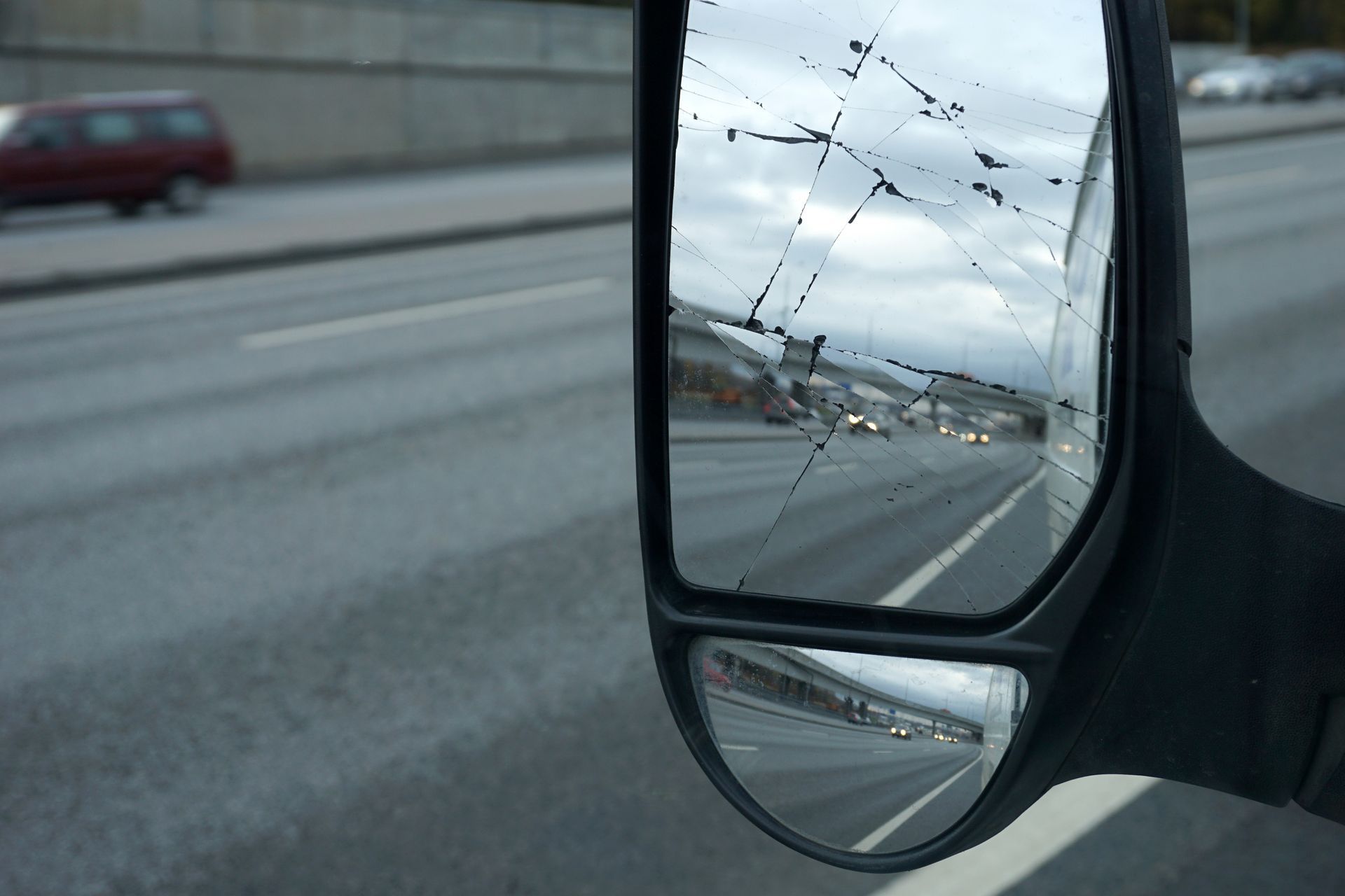 Rétroviseur latéral fissuré reflétant une route mouillée et un ciel nuageux, avec une voiture rouge floue en arrière-plan.