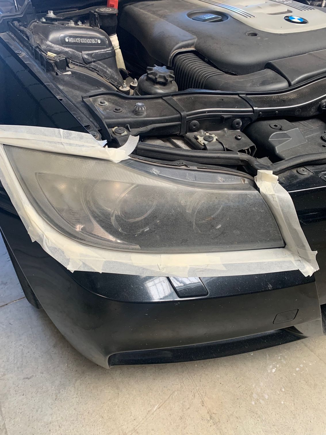 Gros plan avant gauche d'une voiture gris foncé avec un phare détaché et éraflé et un pare-chocs endommagé dans un garage.