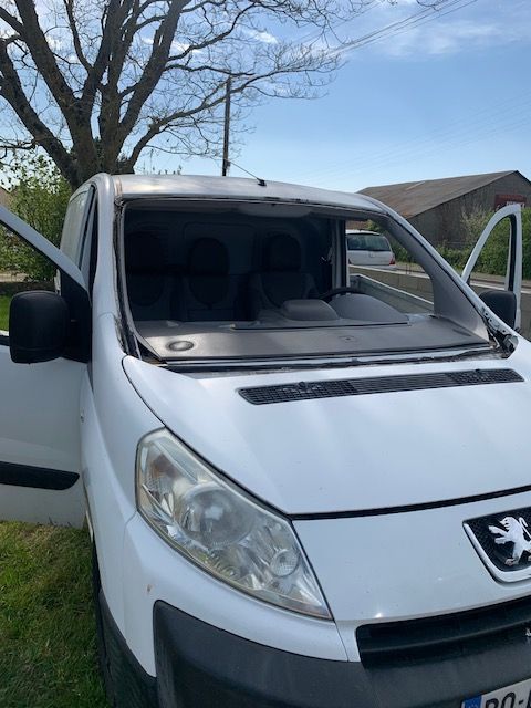 Une camionnette Peugeot blanche avec un pare-brise cassé est garée à l'extérieur, près d'un arbre et d'une maison.