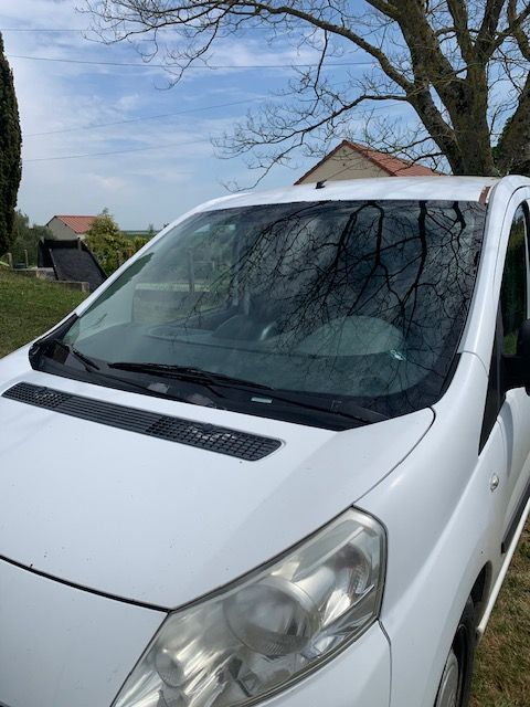 Une camionnette blanche garée à l'extérieur sous un ciel bleu, avec des arbres et des maisons se reflétant dans le pare-brise.