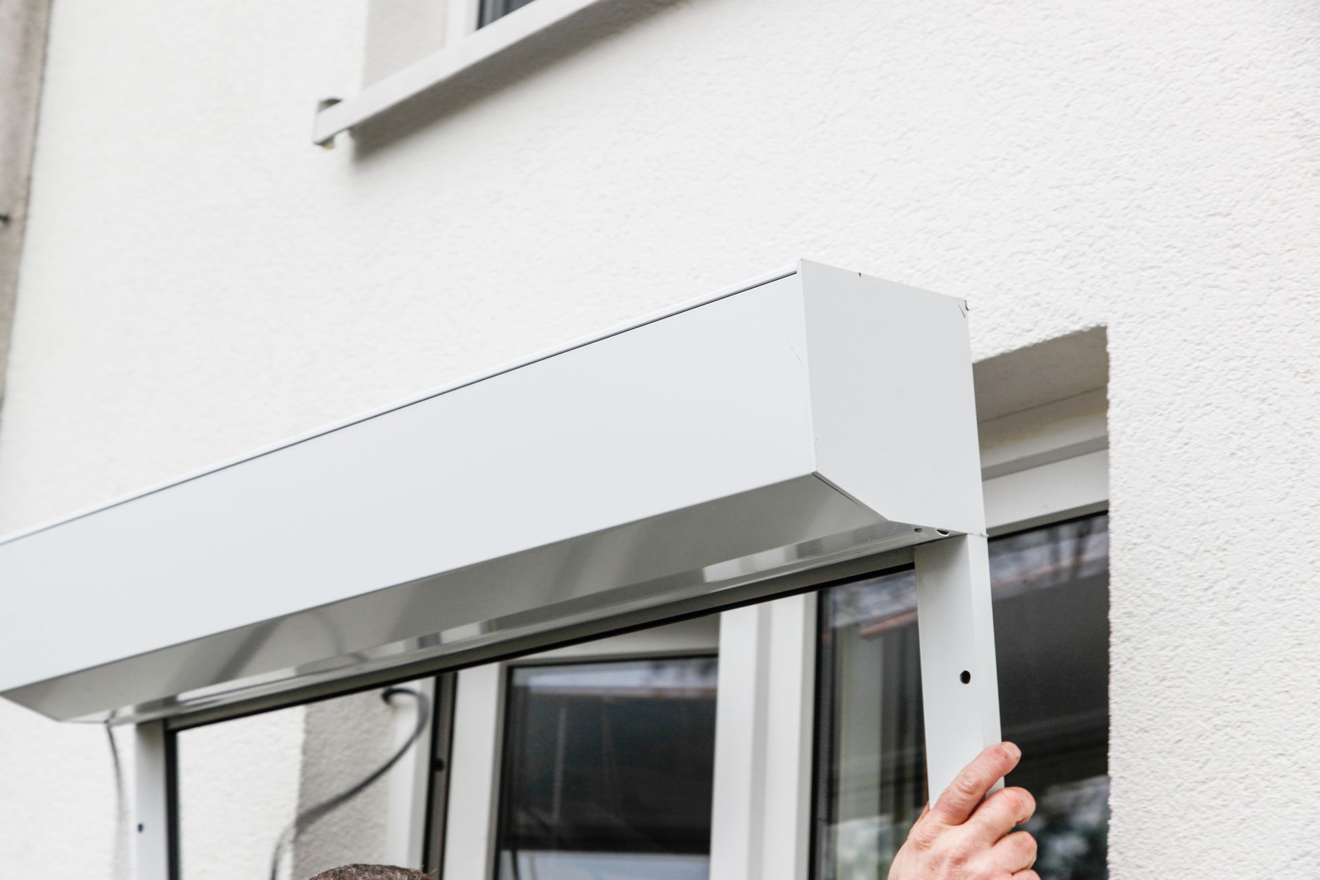Un store blanc recouvre une fenêtre tandis qu'une main en ajuste le bord sur la façade d'un bâtiment.