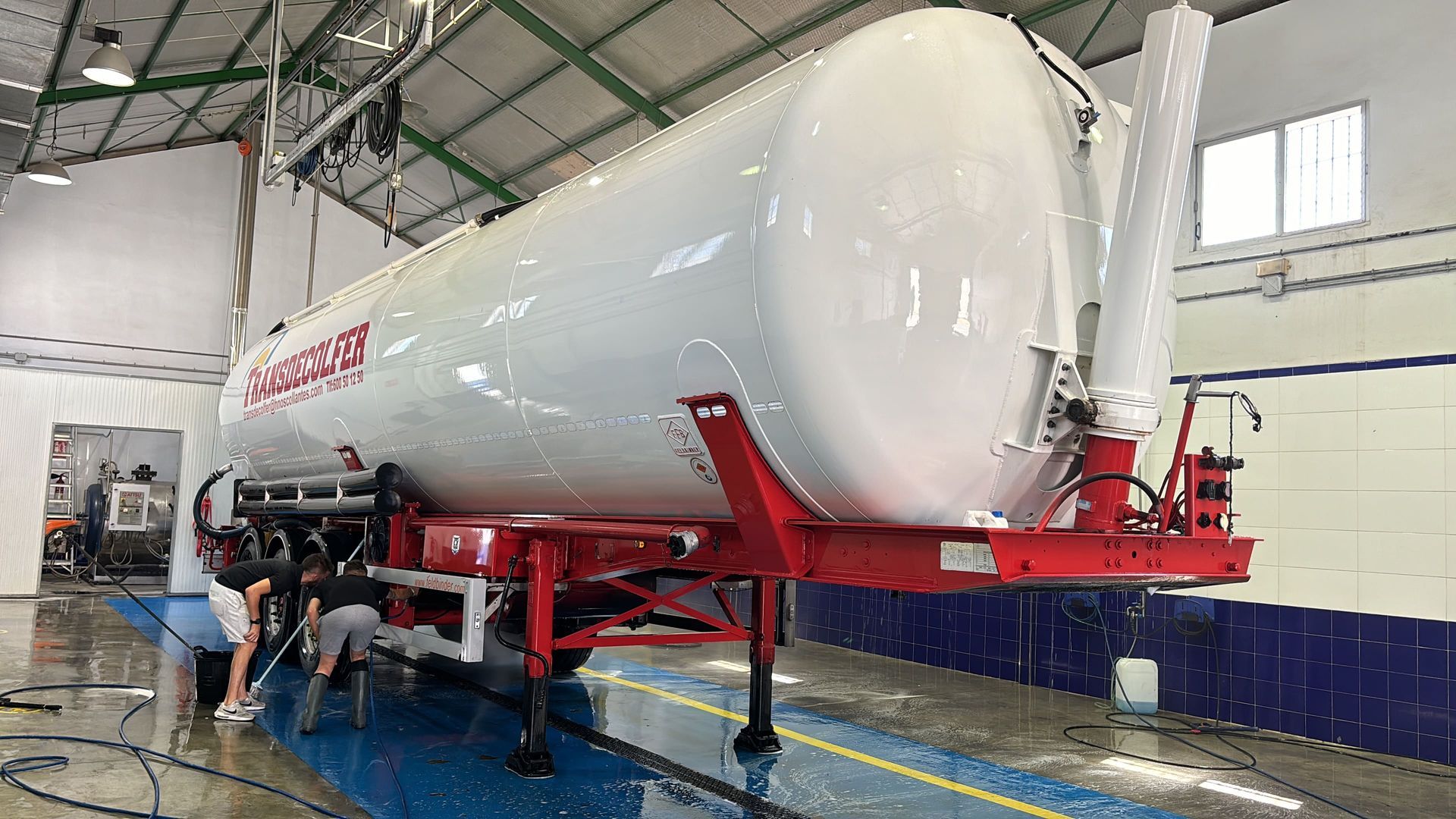 Un gran camión cisterna blanco sobre un marco rojo se lava en un muelle; dos personas lo están lavando con manguera.