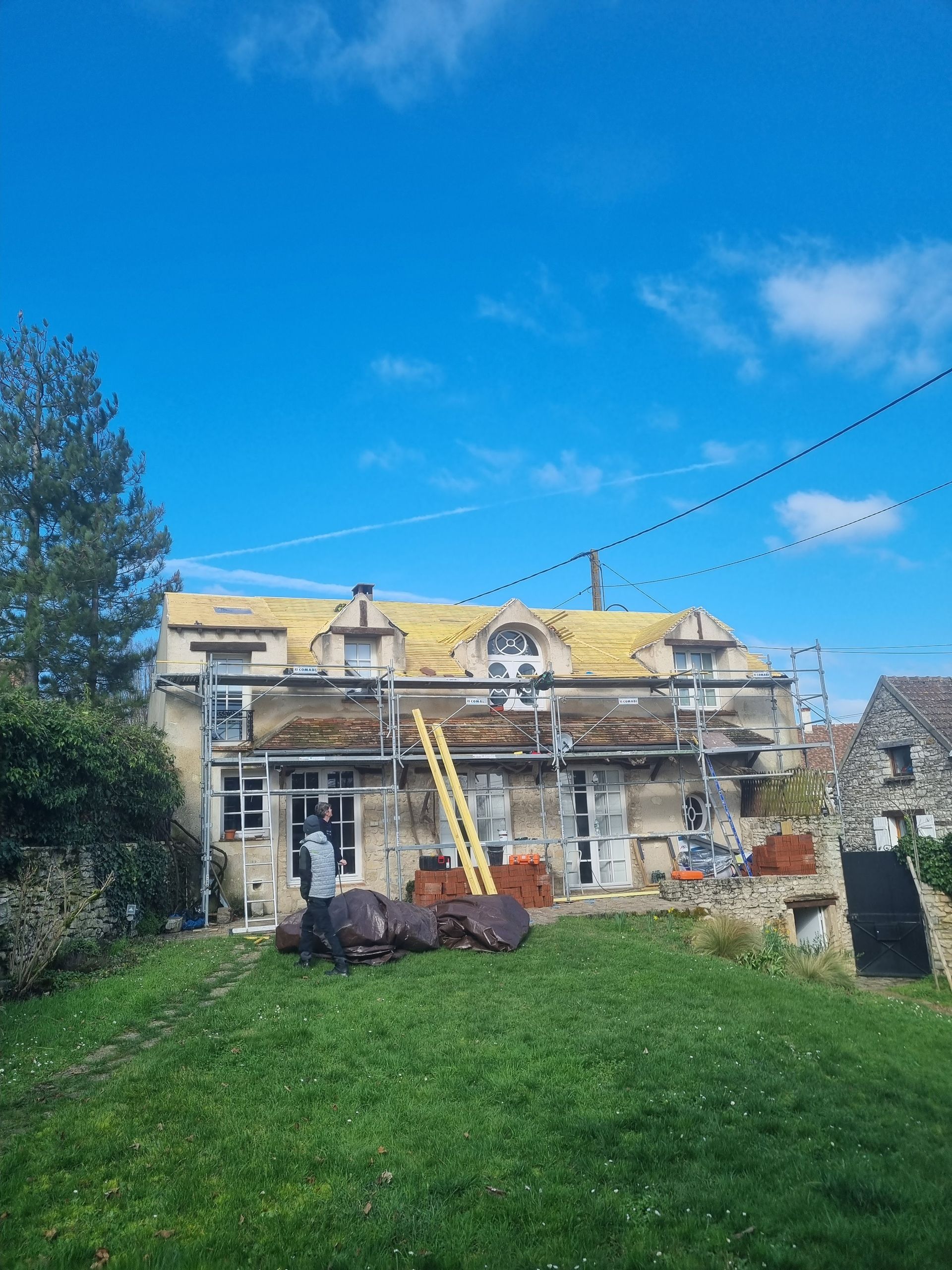 Maison en rénovation avec échafaudages, une charpente apparente et une personne près de matériaux sur la pelouse.