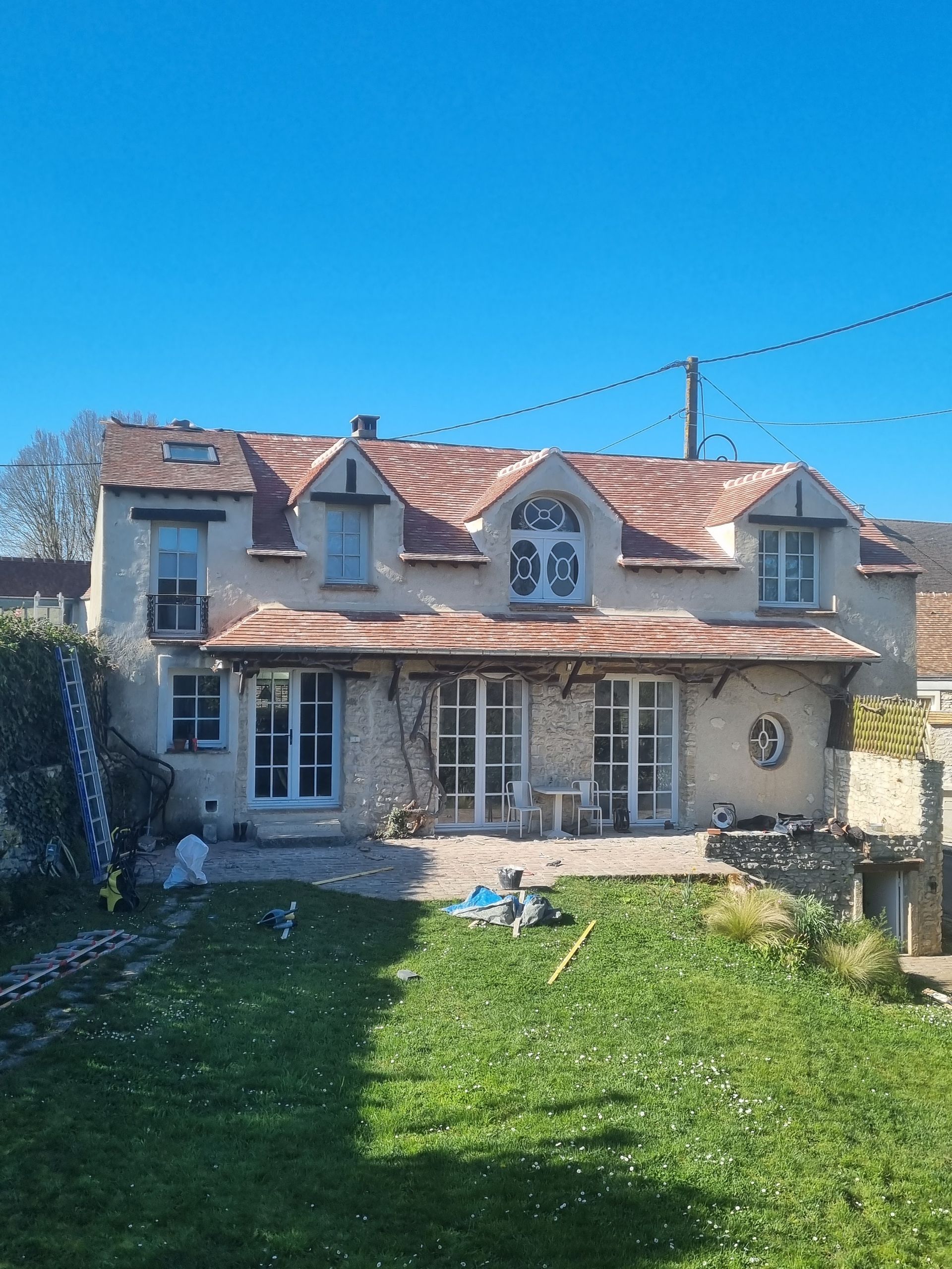 Maison à deux étages avec toit rouge, murs beiges, encadrements de fenêtres blancs et grandes portes vitrées. Pelouse verte, ciel bleu.