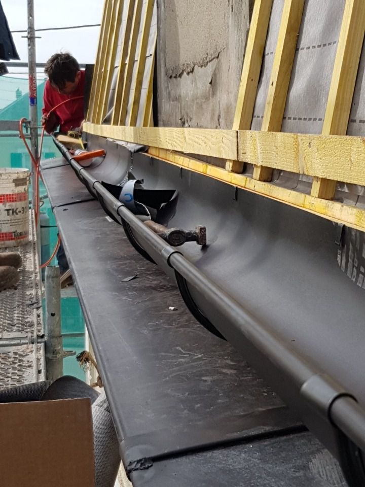 Un homme installe une gouttière sur un toit, avec un échafaudage en arrière-plan. On distingue une gouttière noire, un tuyau gris et des poutres en bois.