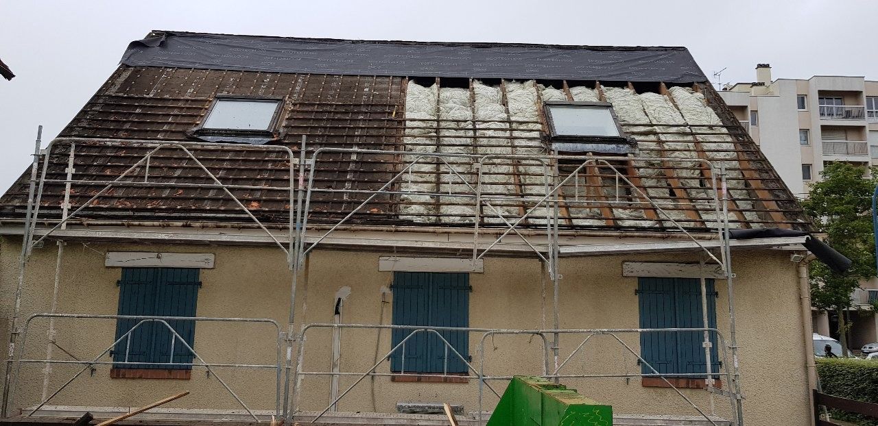 Toiture en cours de rénovation : échafaudages, puits de lumière et bardeaux patinés. Volets bleus au rez-de-chaussée.