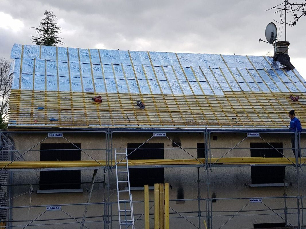 Toiture d'une maison en construction. Échafaudages, poutres en bois, isolant bleu et ouvriers visibles.