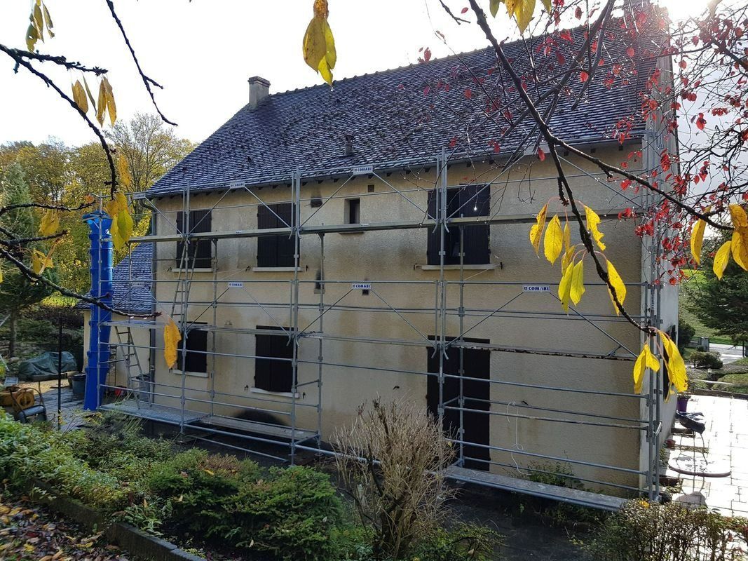 Maison en rénovation avec échafaudages, fenêtres apparentes et toit gris. Feuilles d'automne au premier plan.