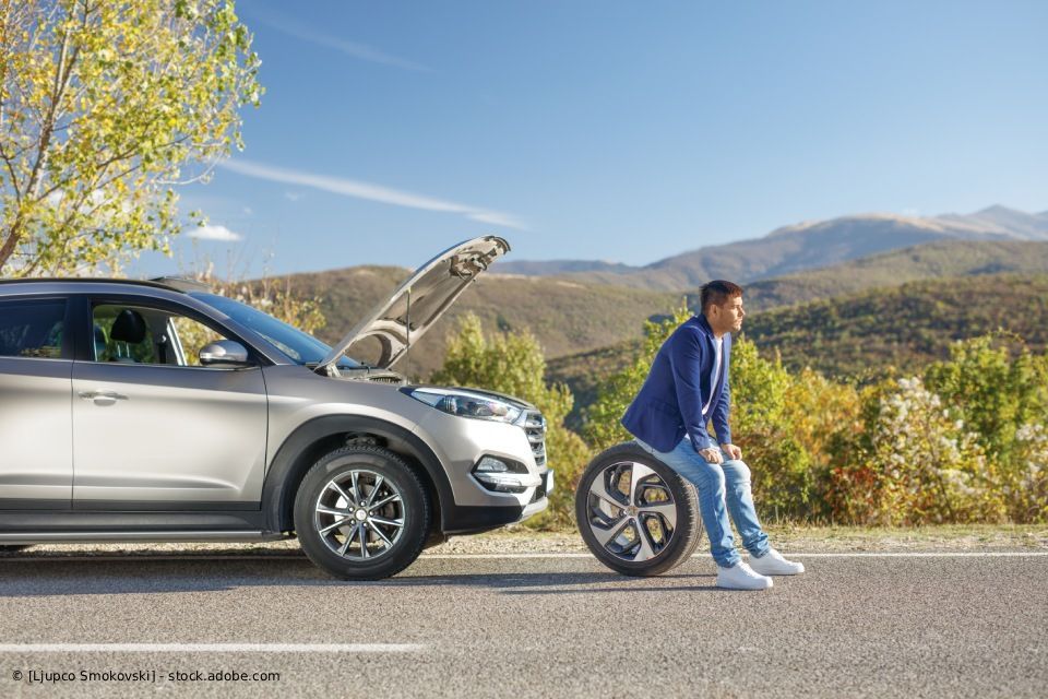 Mann sitzt auf einem Reifen neben einem liegengebliebenen Auto mit geöffneter Motorhaube, im Hintergrund ein Bergpanorama.