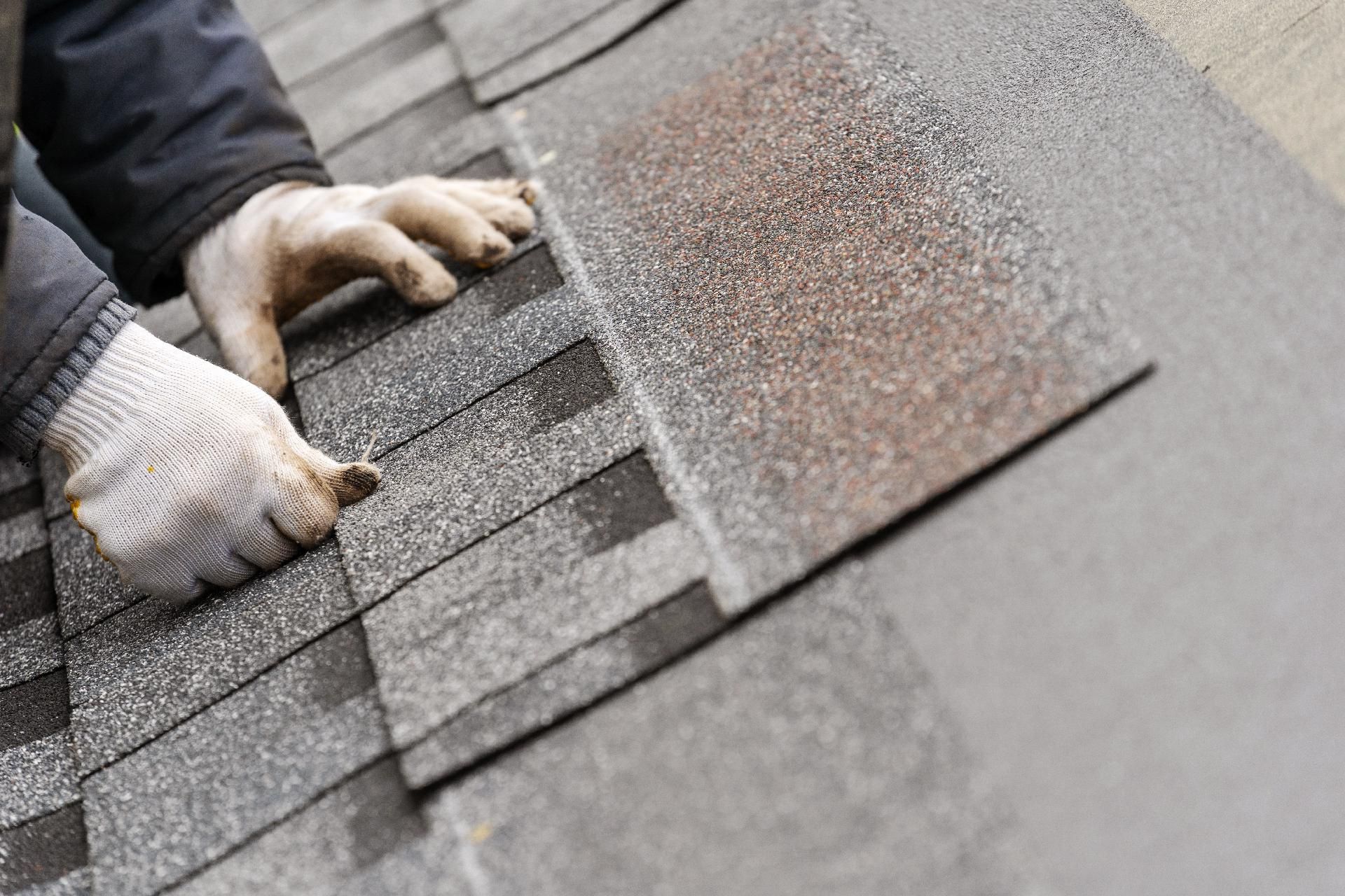 Hände in Arbeitshandschuhen beim Verlegen grauer Asphalt-Dachschindeln.