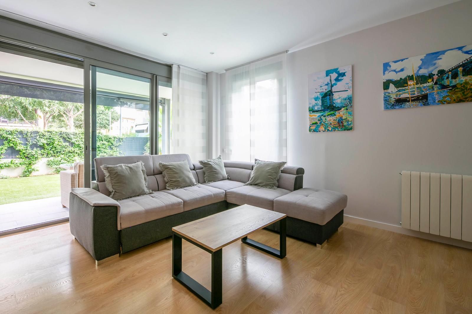 Sala de estar con sofá de color claro, mesa de centro de madera y grandes puertas de vidrio que conducen a un patio verde.