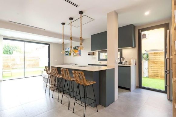 Cocina moderna con isla, asientos y acceso al exterior. Gabinetes grises, encimera beige y abundante luz natural.