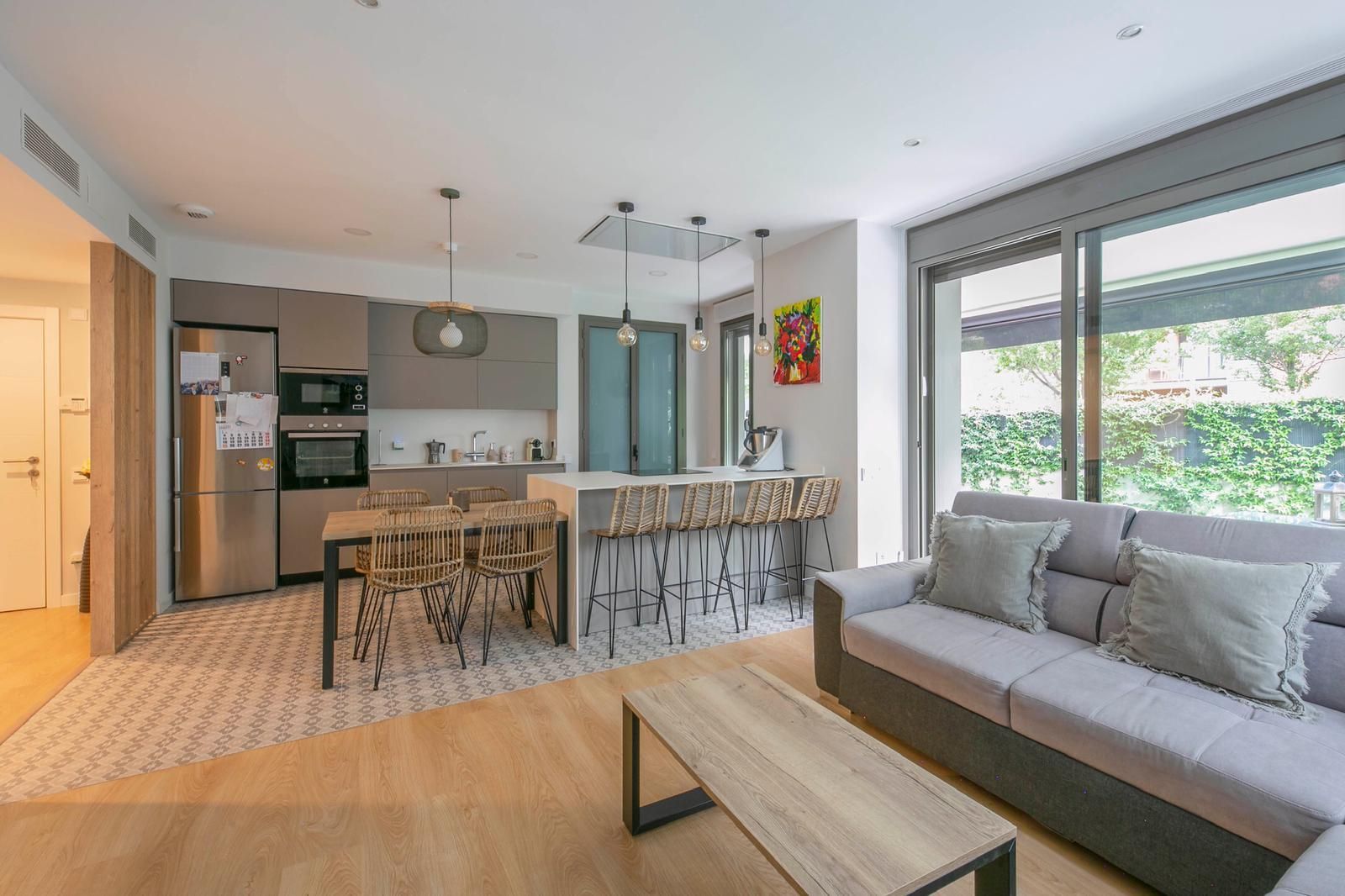 Moderna cocina abierta y sala de estar con electrodomésticos de acero inoxidable, sofá gris claro y mesa de madera.