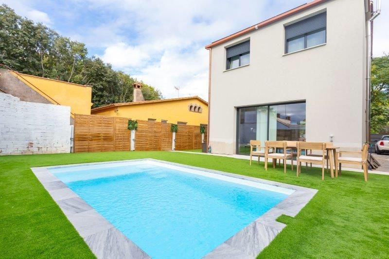 Una casa moderna con piscina y comedor al aire libre sobre un césped verde. Cielo azul.