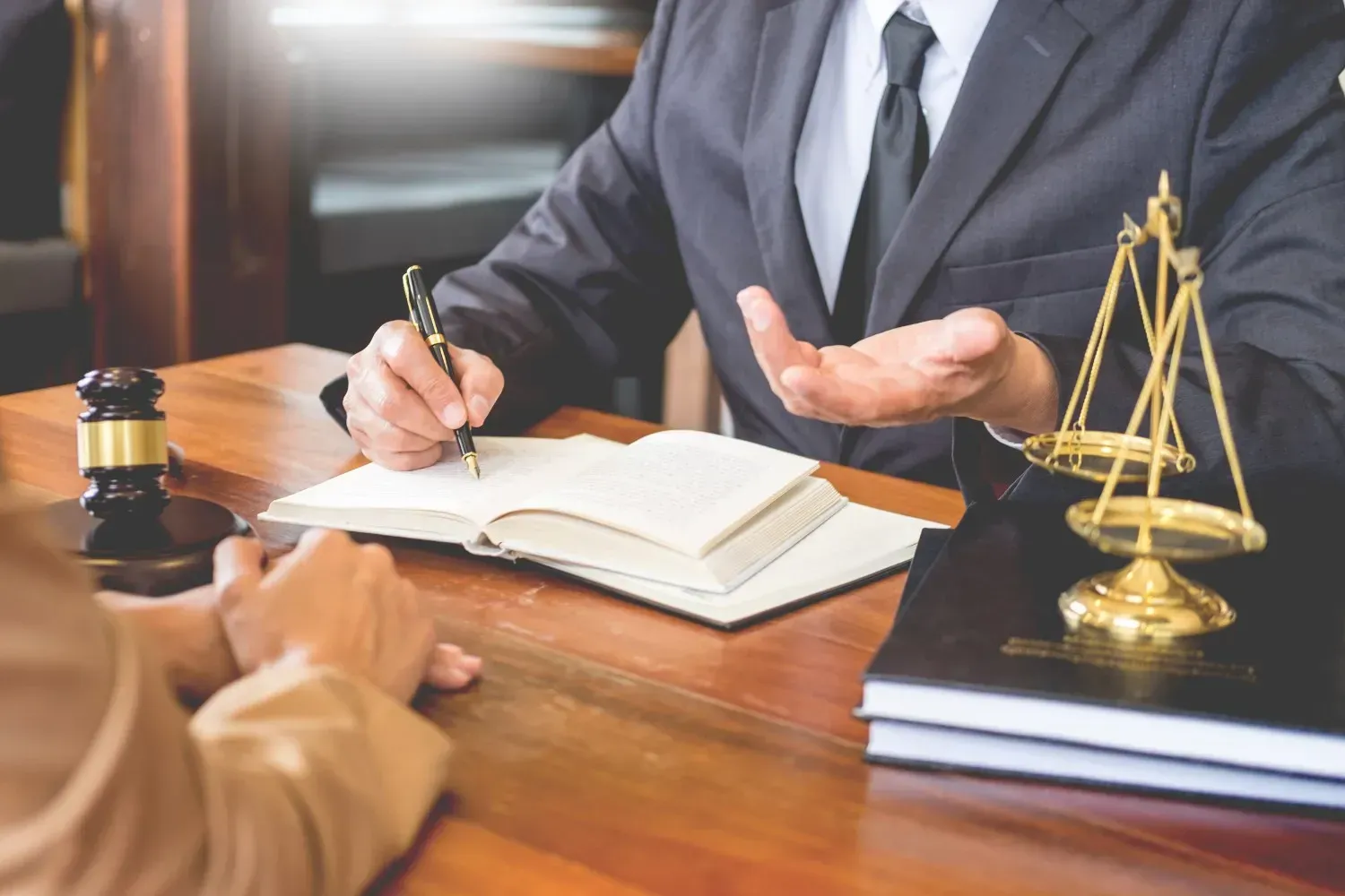 Un profesional vestido de traje está sentado en un escritorio con un mazo y una balanza de la justicia, escribiendo en un libro mientras hace gestos hacia un cliente.