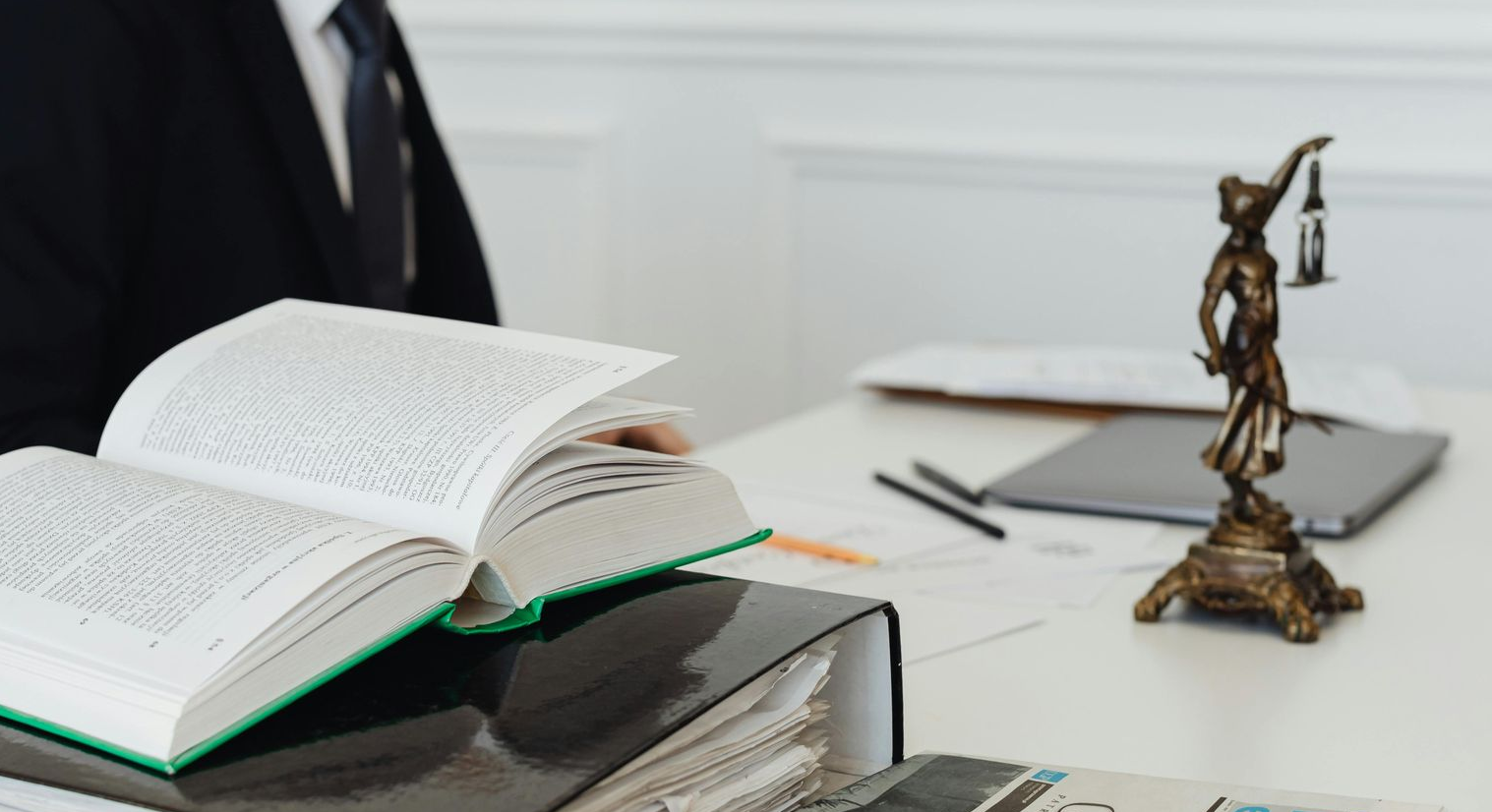 Un profesional vestido de traje está sentado en un escritorio con un libro de leyes abierto y una estatua de bronce de la Dama de la Justicia.