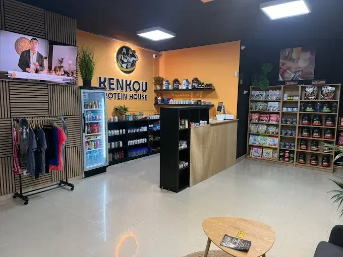 Interior de una tienda de suplementos con estantes de productos, un mostrador y un televisor.