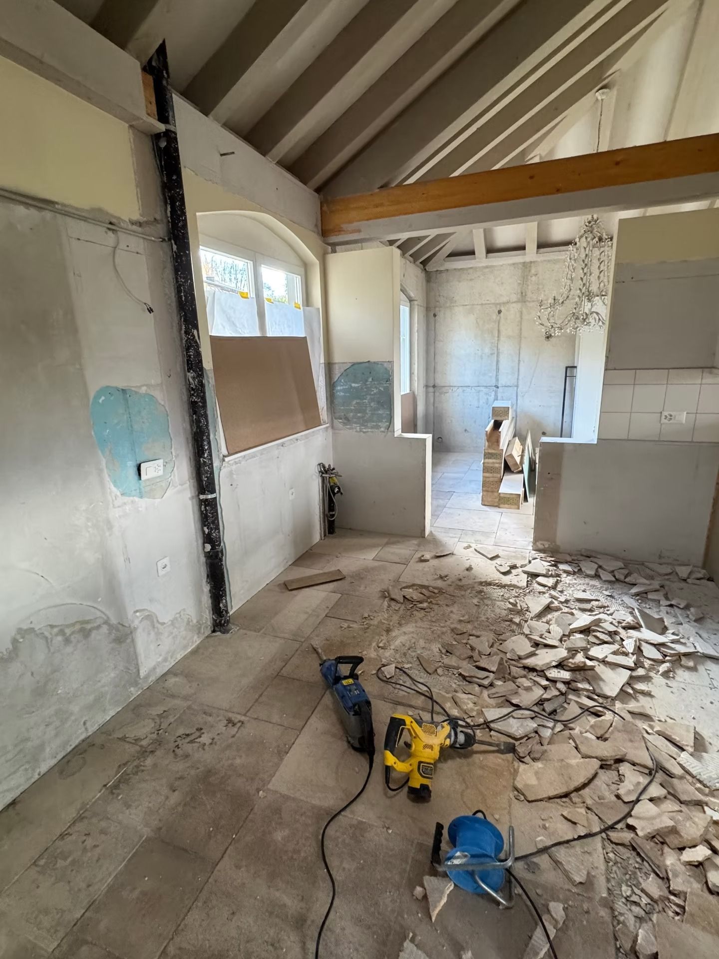 Pièce en rénovation avec poutres apparentes, plâtre cassé au sol et outils électriques près d'une fenêtre.