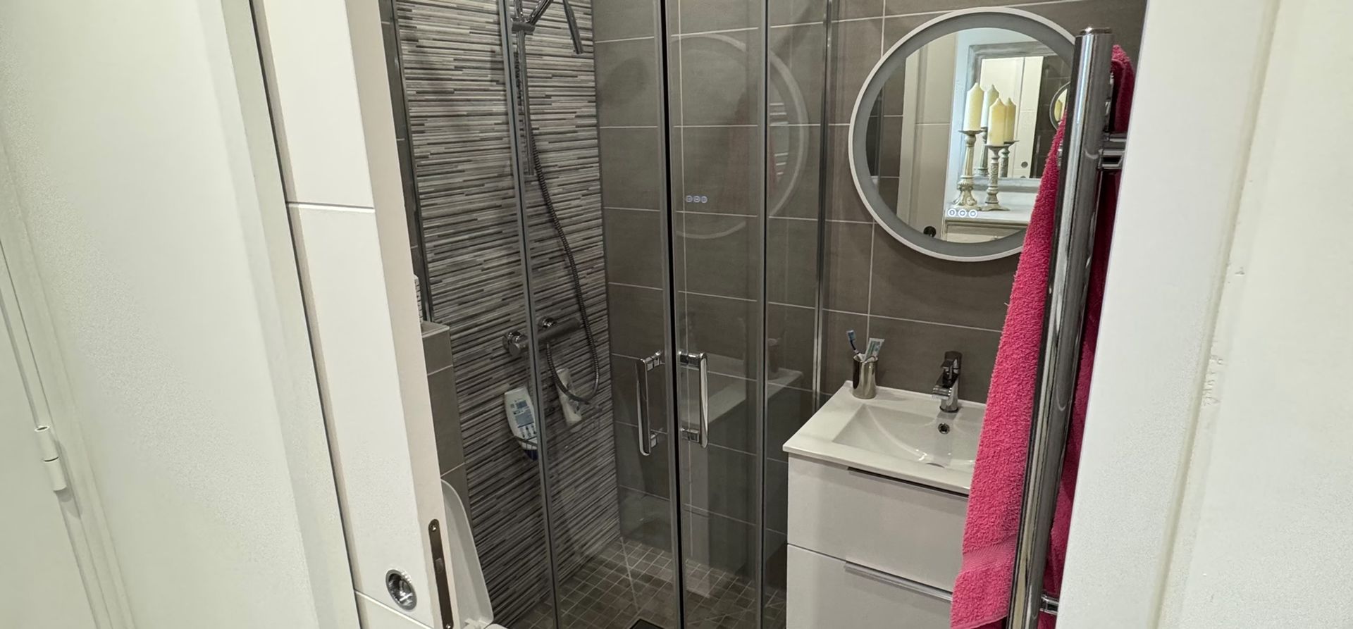 Petite salle de bains avec douche en verre, miroir rond, lavabo blanc et serviettes rayées suspendues sur les côtés.