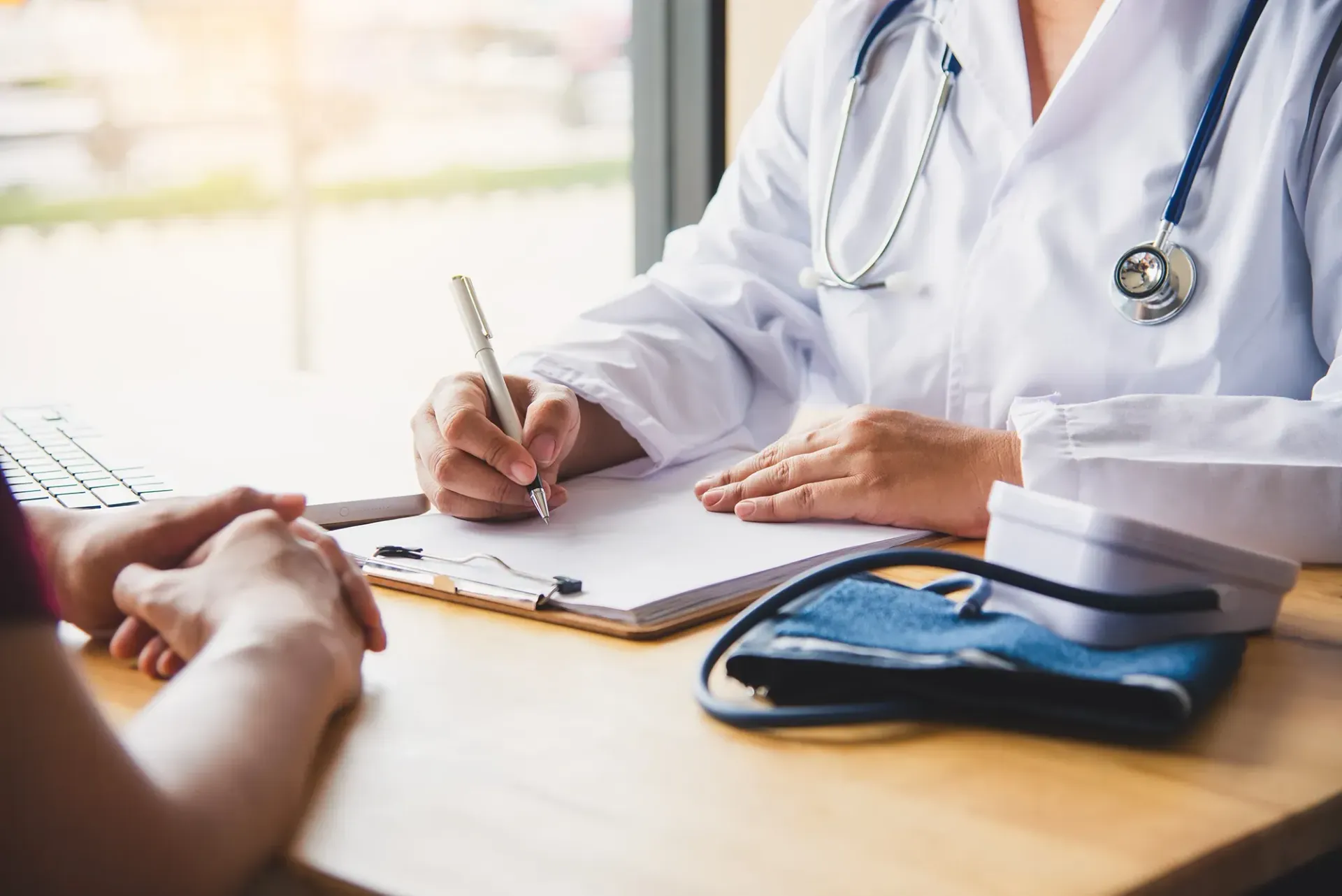 Ein Arzt sitzt mit einem Patienten an einem Tisch und schreibt auf ein Klemmbrett.