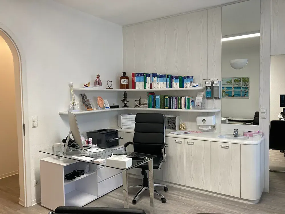Eine Arztpraxis mit Schreibtisch, Stuhl, Computer und Regalen.