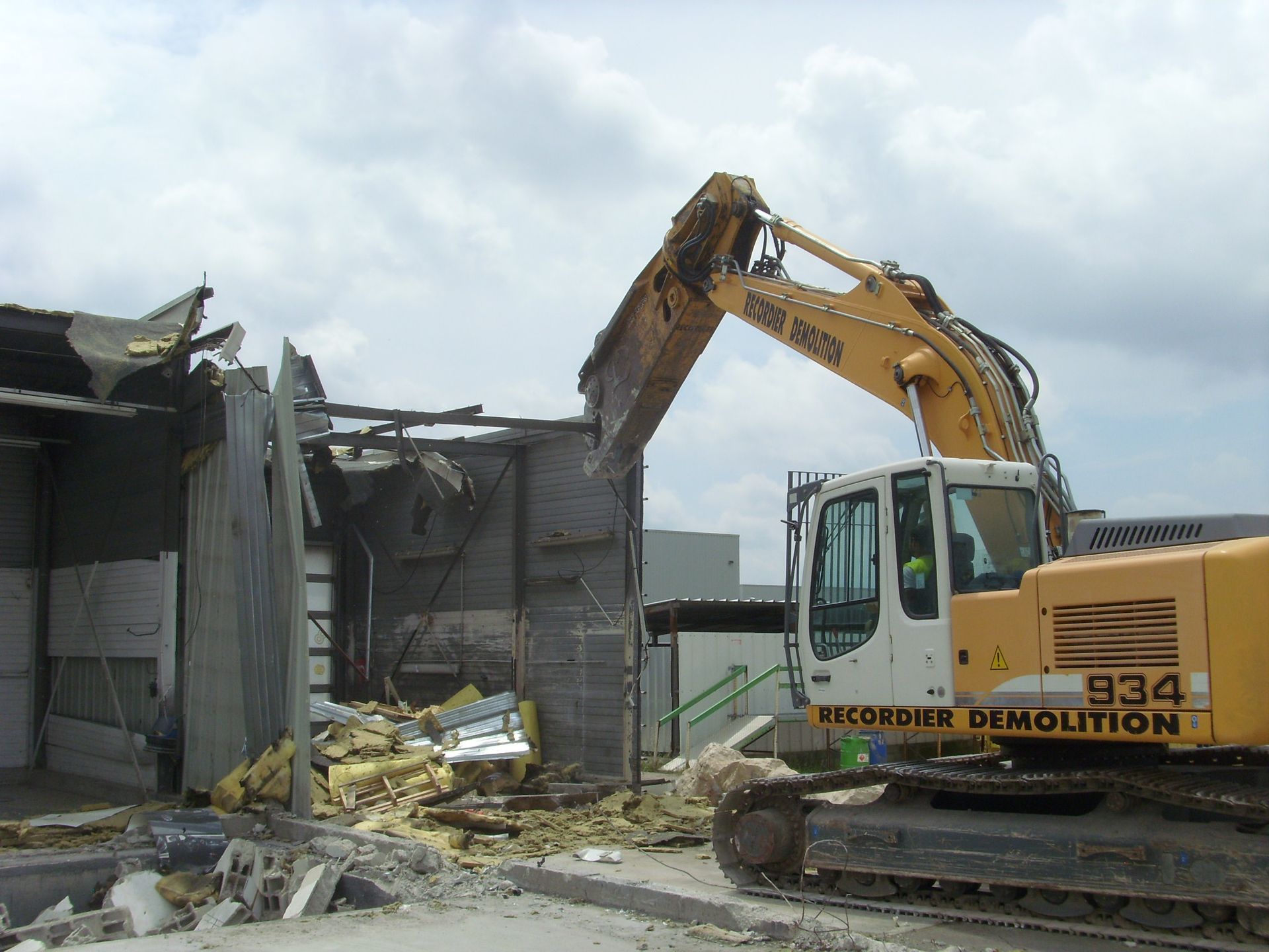 Une pelleteuse jaune démolit un bâtiment ; débris et structure arrachée sont visibles. Ciel nuageux.