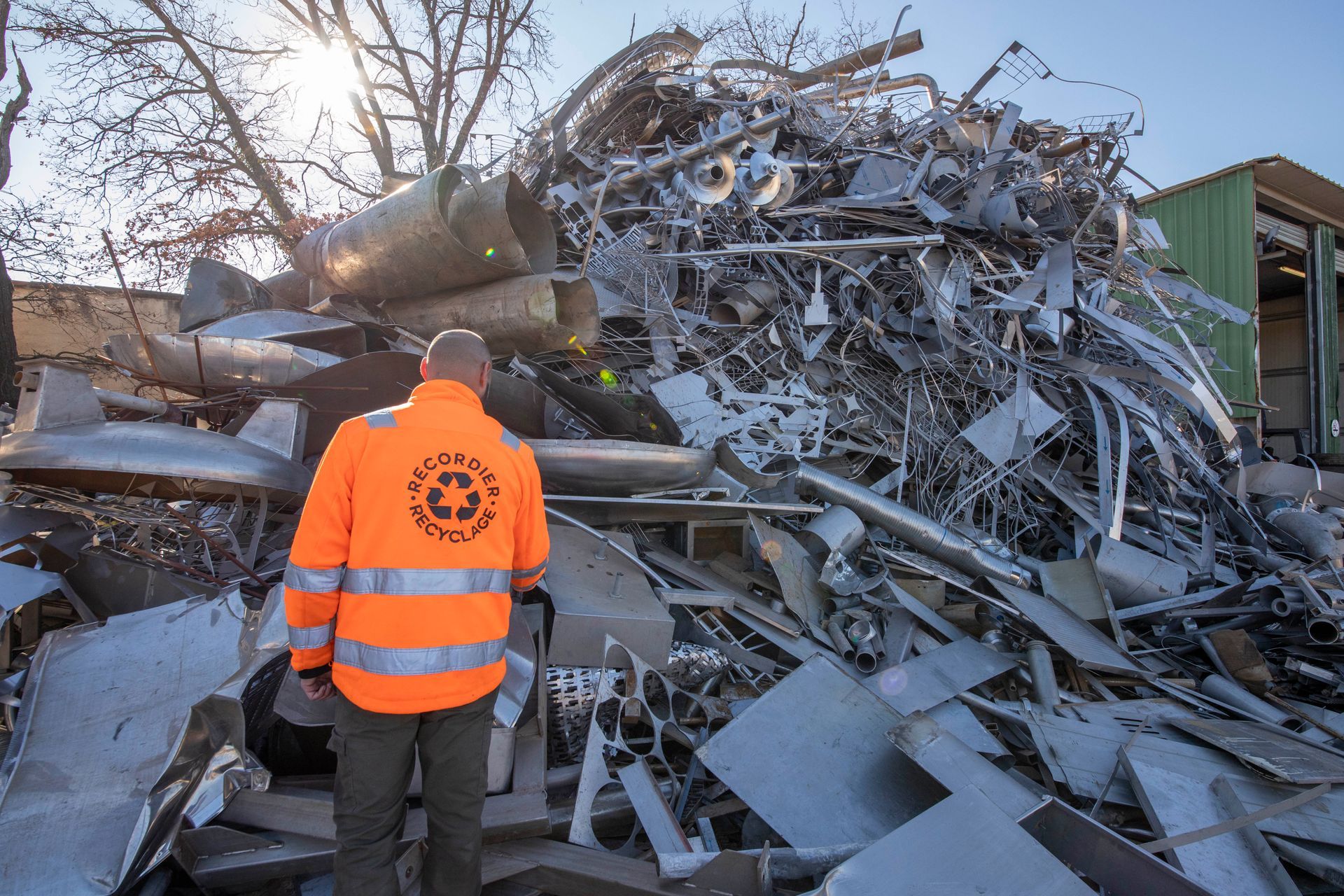 Un employé vêtu d'un gilet orange se tient devant un grand tas de ferraille à l'extérieur.