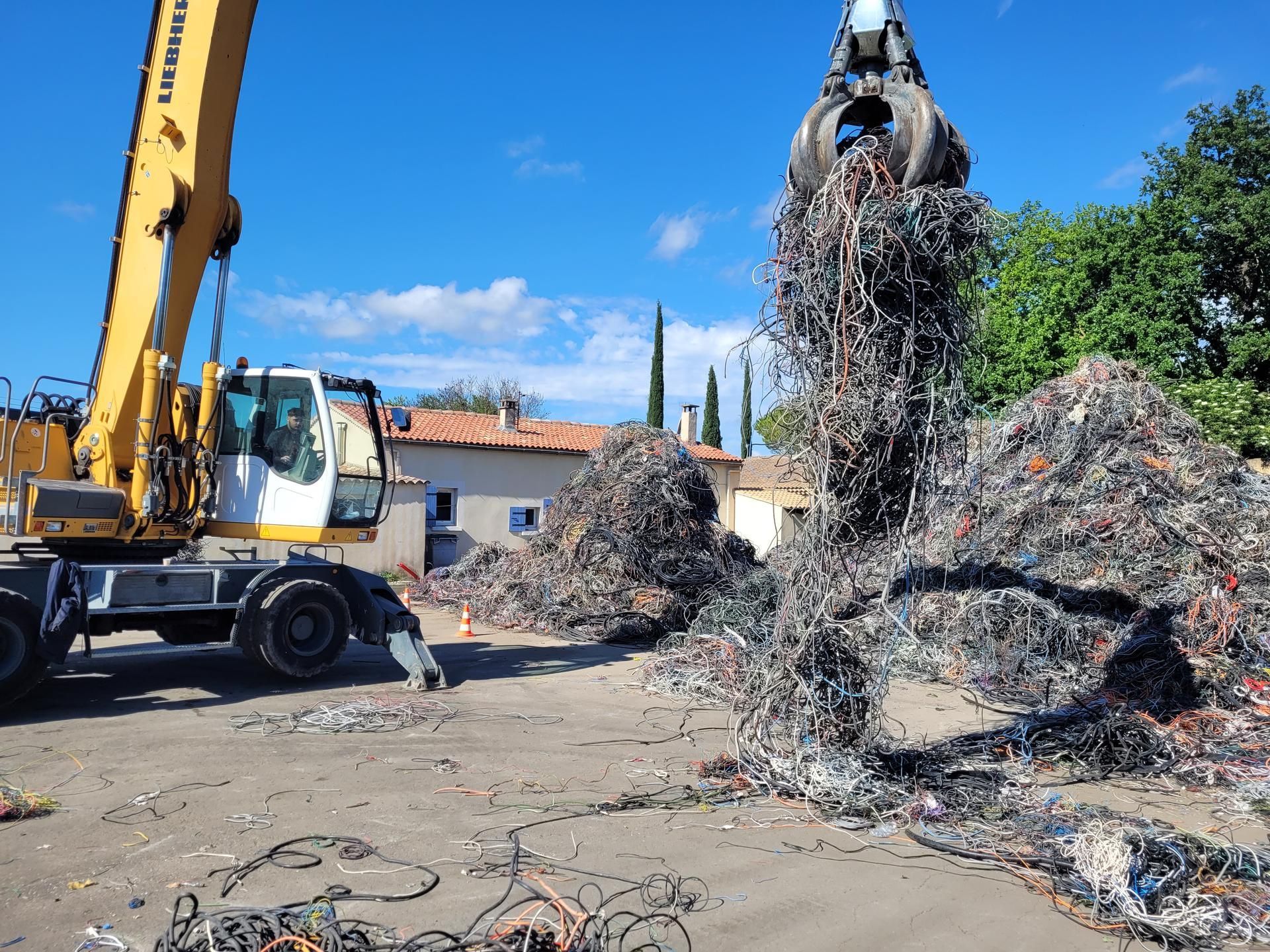 Une excavatrice à griffes soulève de la ferraille dans le centre de recyclage ETS ANDRE RECORDIER sous un ciel bleu.