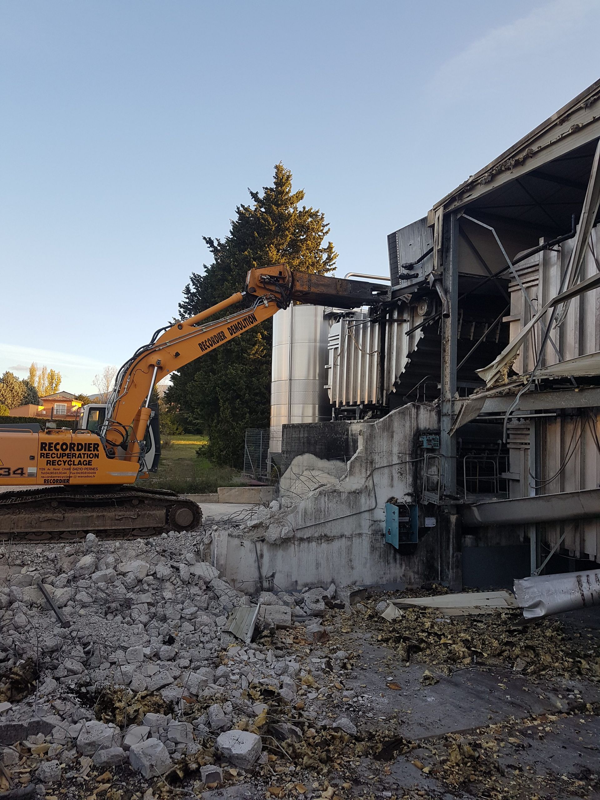 Une excavatrice démolit un bâtiment, révélant un silo métallique ; des débris de béton sont visibles.
