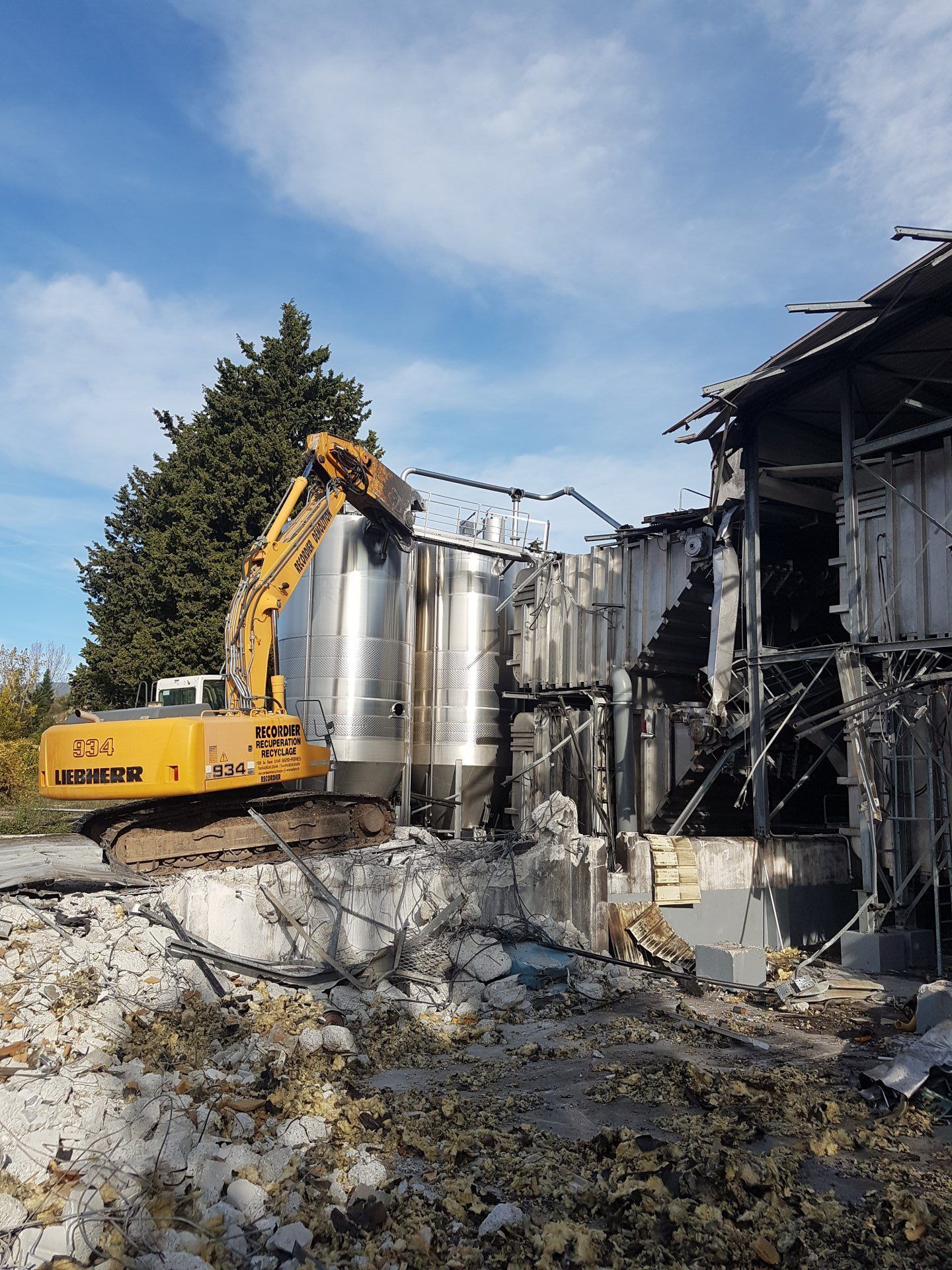 Sous un ciel partiellement nuageux, une pelleteuse jaune démolit un bâtiment endommagé abritant des réservoirs en acier inoxydable.