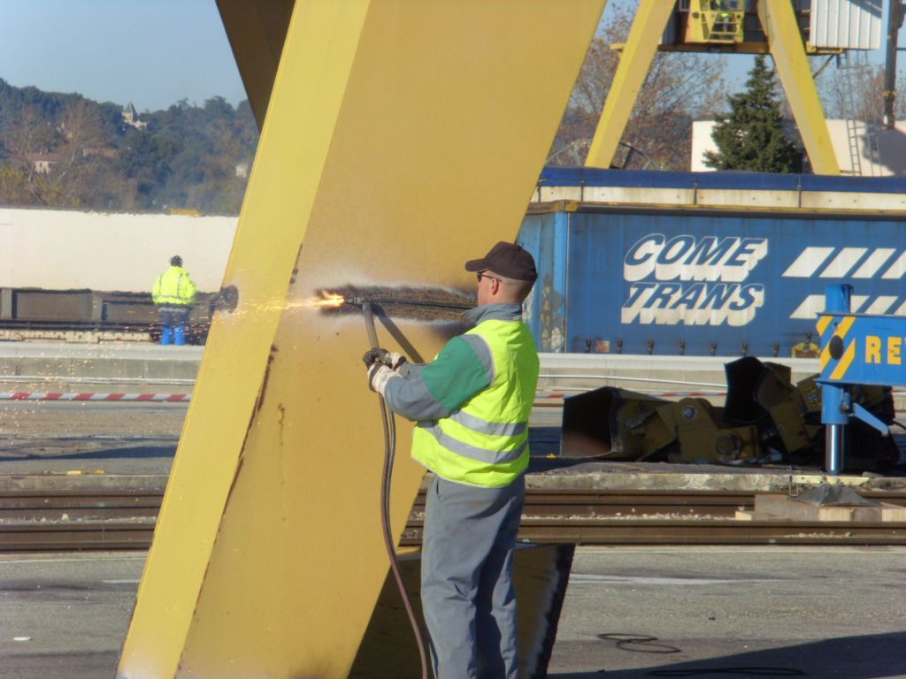Un ouvrier utilise une torche sur une structure métallique jaune, près d'un conteneur maritime et de voies ferrées.