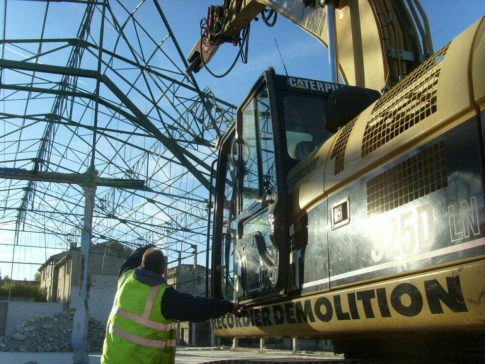 Un ouvrier et une pelleteuse jaune démantèlent un bâtiment à ossature métallique.