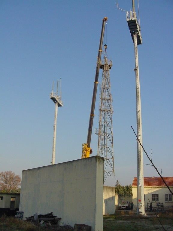 Une grue soulève un élément métallique d'une tour près de deux lampadaires. Ciel bleu, bâtiment beige en arrière-plan.