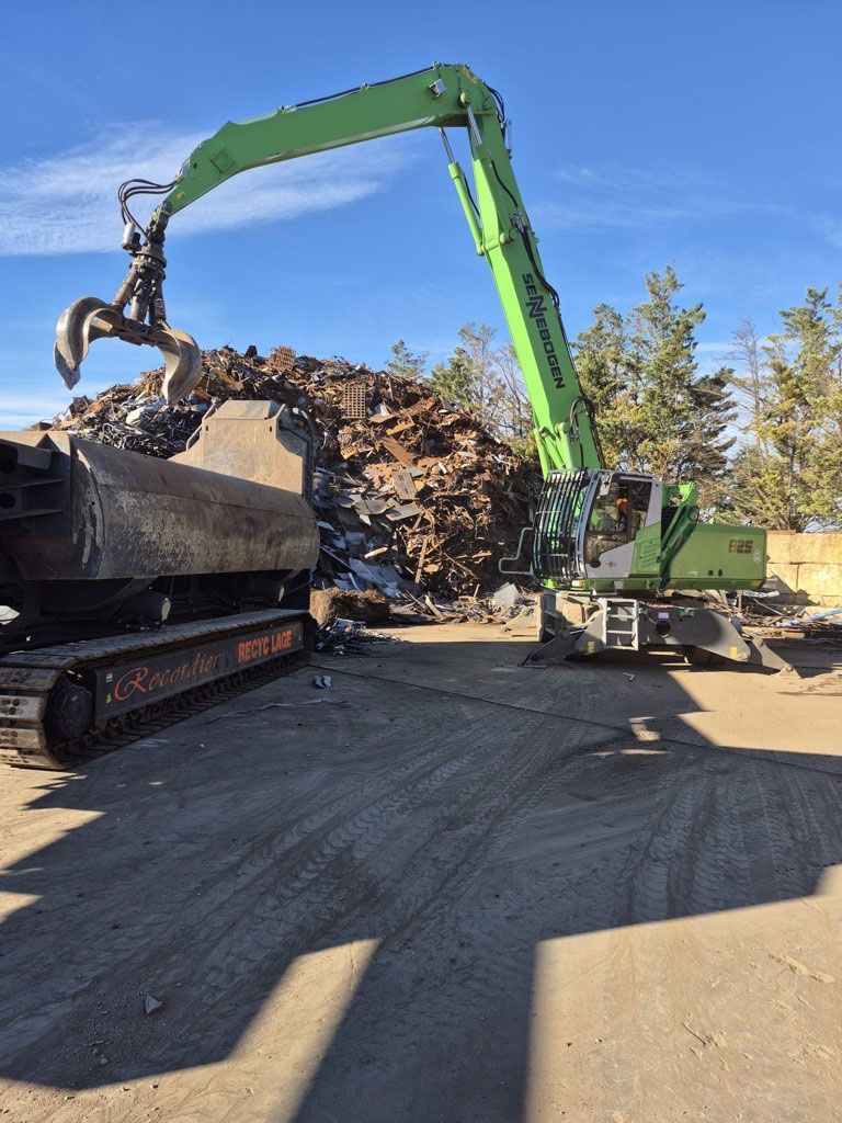 Une pelleteuse verte soulève de la ferraille dans une casse par une journée ensoleillée.