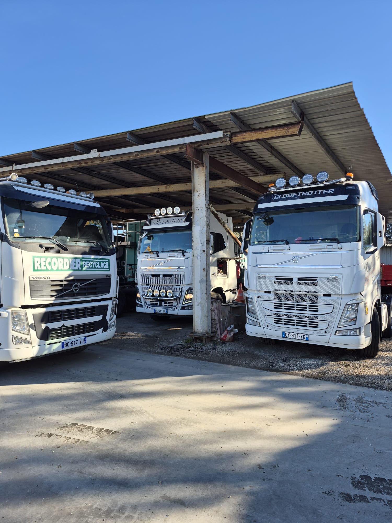 Trois semi-remorques blancs des ETS ANDRE RECORDIER sous un abri métallique ; journée ensoleillée.